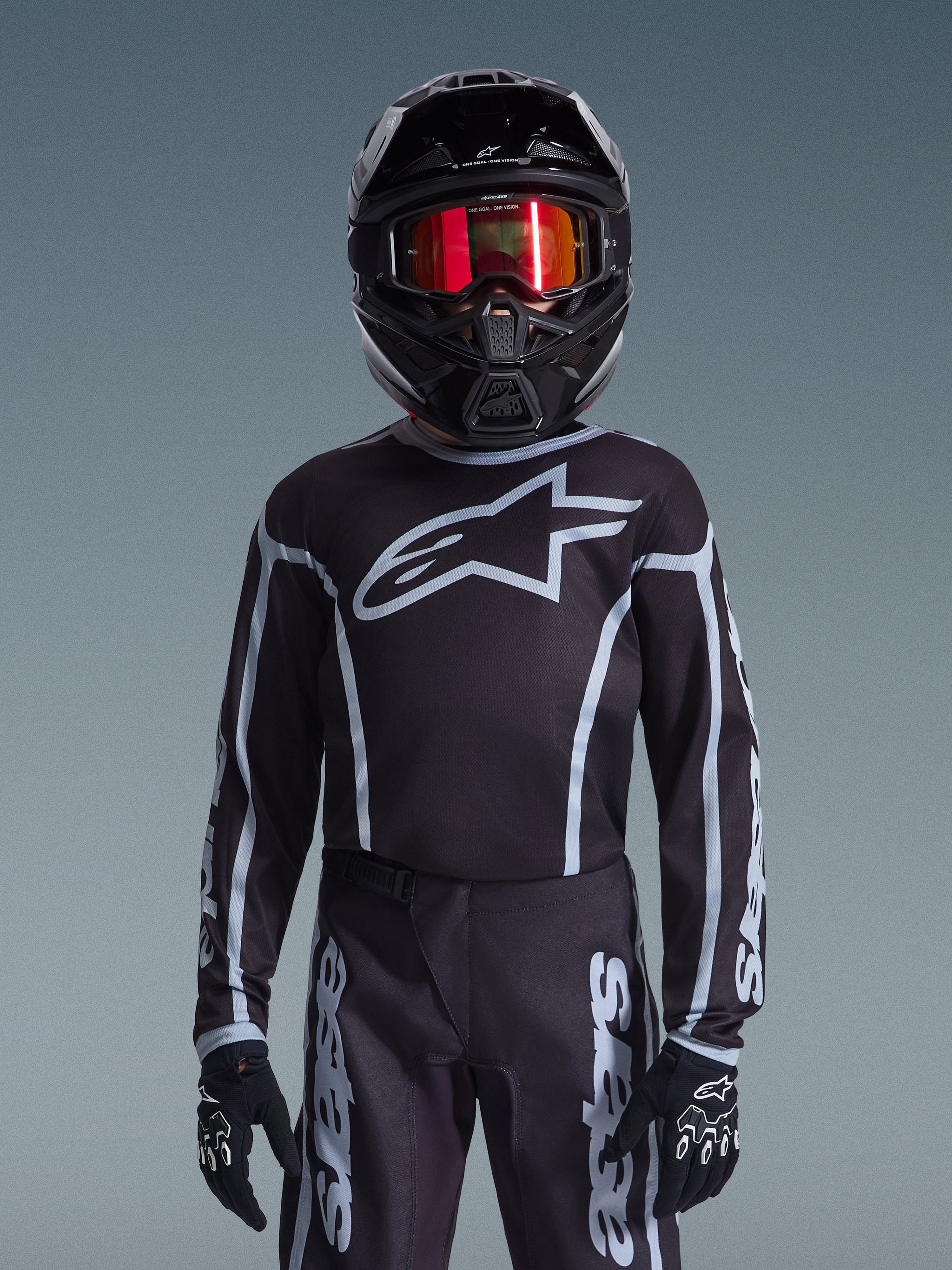 Jeune pilote de motocross portant le maillot Alpinestars 2026 Youth Fluid Apex en noir et gris, pantalon assorti, casque noir avec lunettes réfléchissantes rouges et gants noirs, équipement de course tout-terrain coordonné.