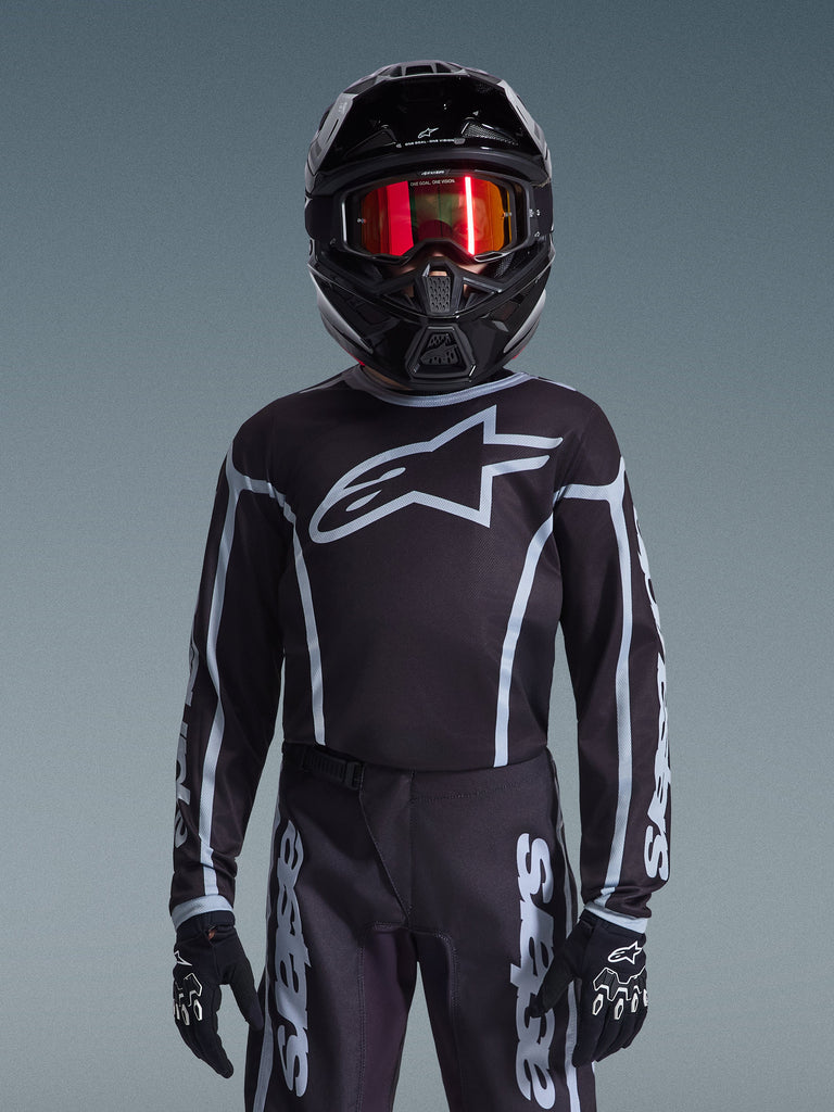 Jeune pilote de motocross portant le maillot Alpinestars 2026 Youth Fluid Apex en noir et gris, pantalon assorti, casque noir avec lunettes réfléchissantes rouges et gants noirs, équipement de course tout-terrain coordonné.