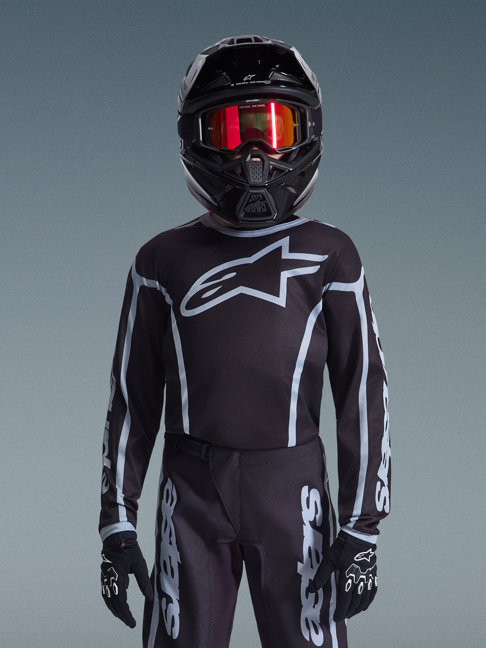 Jeune pilote de motocross portant le maillot Alpinestars 2026 Youth Fluid Apex en noir et gris, pantalon assorti, casque noir avec lunettes réfléchissantes rouges et gants noirs, équipement de course tout-terrain coordonné.