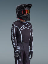 Maillot Junior Alpinestars Fluid Apex 2026 en noir et gris, porté par un jeune pilote avec un casque noir assorti et des lunettes teintées, présentant le logo Alpinestars sur la poitrine et les manches, faisant partie d'un ensemble d'équipement de motocross coordonné.