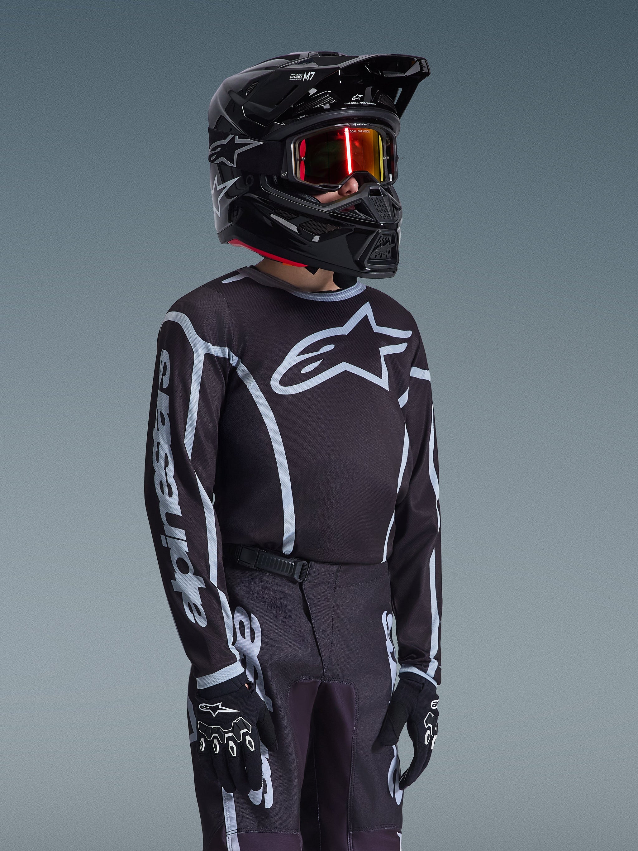 Maillot Junior Alpinestars Fluid Apex 2026 en noir et gris, porté par un jeune pilote avec un casque noir assorti et des lunettes teintées, présentant le logo Alpinestars sur la poitrine et les manches, faisant partie d'un ensemble d'équipement de motocross coordonné.