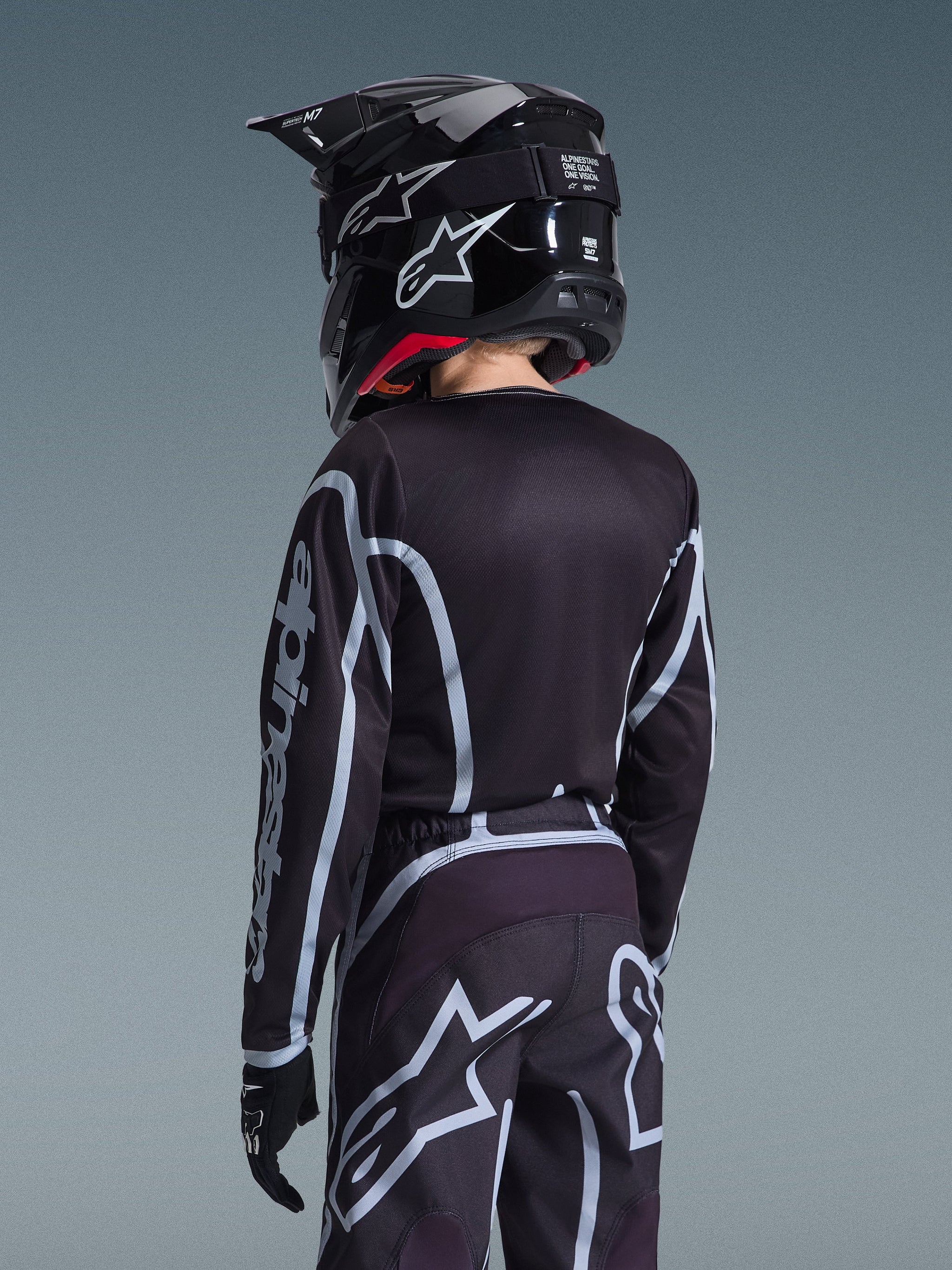 Vue arrière d'un jeune pilote de motocross portant le maillot Alpinestars 2026 Youth Fluid Apex et le pantalon assorti en noir et gris, avec un casque noir arborant des logos blancs et des gants noirs, équipement de course tout-terrain athlétique pour jeunes pilotes.