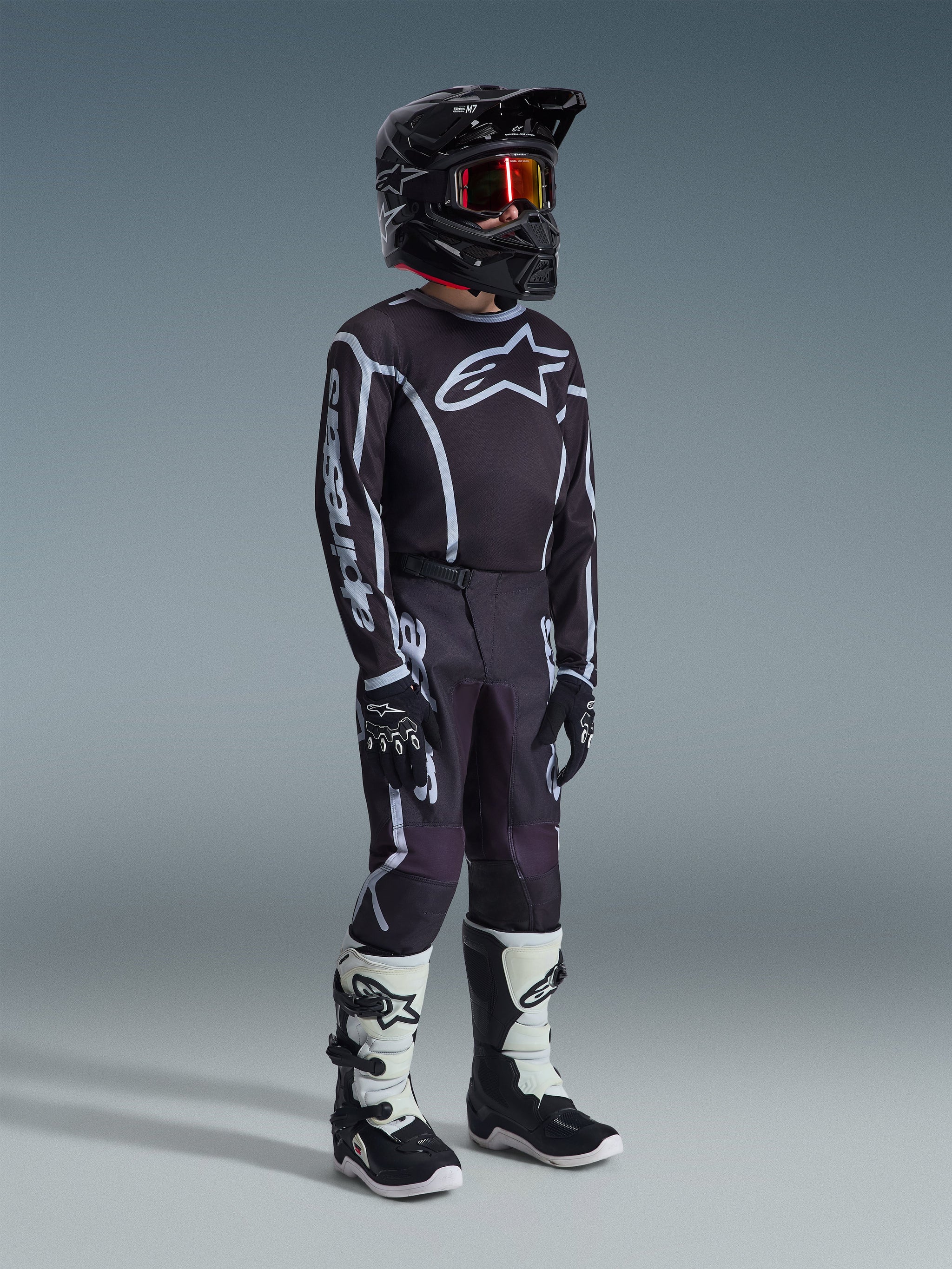 Jeune pilote de motocross portant le maillot Alpinestars Youth Fluid Apex 2026 en Noir Gris, pantalon noir et gris assorti avec graphismes blancs, casque noir avec lunettes teintées rouges, gants noirs et bottes de motocross blanches et noires, tenue complète de course tout-terrain.