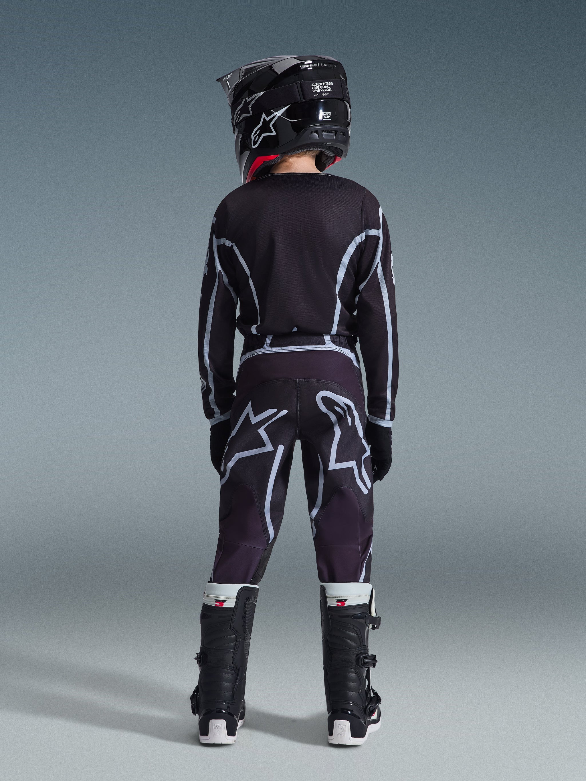 Alpinestars Maillot Junior Fluid Apex 2026, jeune pilote de motocross vu de dos, maillot noir et gris, pantalon assorti avec larges logos blancs, casque noir et bottes Tech Series noir et blanc, tenue de performance complète pour le tout-terrain.
