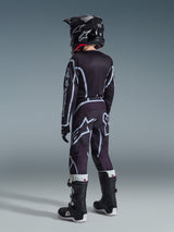 Maillot Alpinestars Youth Fluid Apex 2026, maillot de motocross, noir et gris, porté avec le pantalon Youth Fluid Grid assorti, un casque noir et des bottes noires et blanches, vue arrière montrant une tenue complète de motocross pour jeunes pilotes