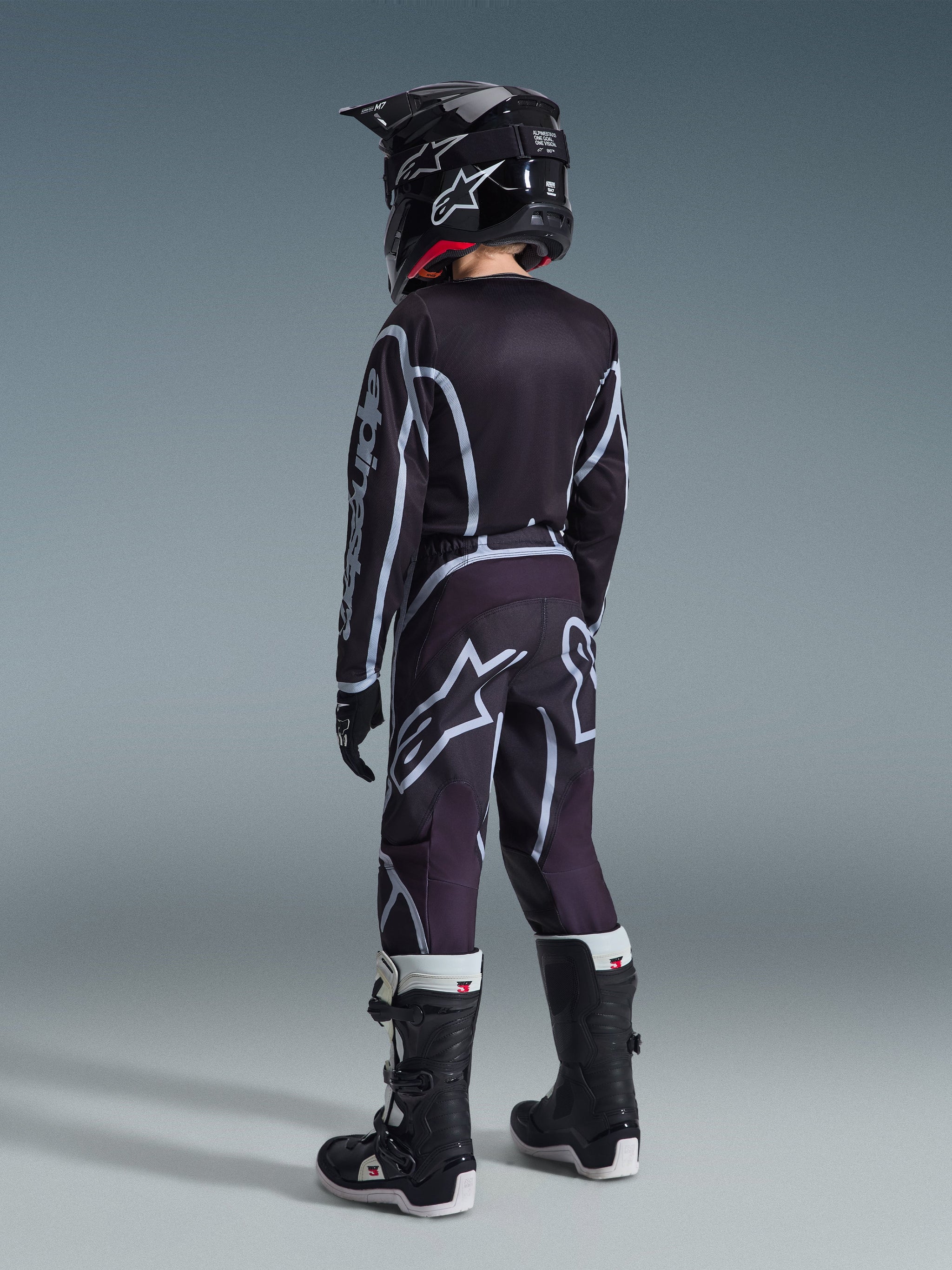 Maillot Alpinestars Youth Fluid Apex 2026, maillot de motocross, noir et gris, porté avec le pantalon Youth Fluid Grid assorti, un casque noir et des bottes noires et blanches, vue arrière montrant une tenue complète de motocross pour jeunes pilotes