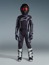 Jeune pilote de motocross portant le maillot Alpinestars Youth Fluid Apex 2026 noir et gris, avec pantalon assorti, casque noir et lunettes rouges irisées, gants noirs et blancs et bottes de motocross, tenue complète pour la course tout-terrain junior