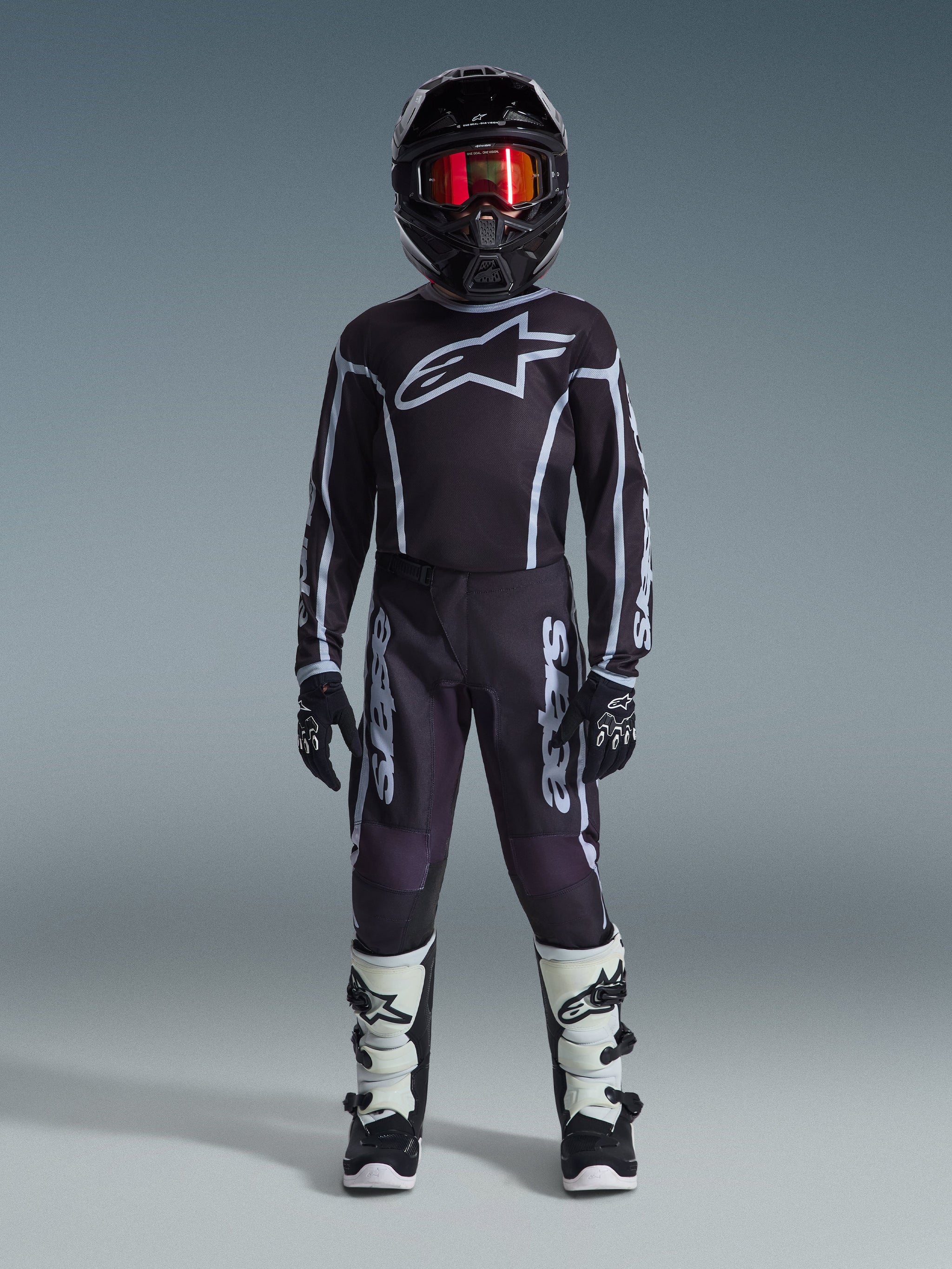 Jeune pilote de motocross portant le maillot Alpinestars Youth Fluid Apex 2026 noir et gris, avec pantalon assorti, casque noir et lunettes rouges irisées, gants noirs et blancs et bottes de motocross, tenue complète pour la course tout-terrain junior