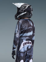 Youth rider wearing complete motocross gear from behind, Alpinestars 2026 Youth Fluid Lahnd Jersey in Iron Camo with black and light blue patterns, matching pants with 'astars' logo on the seat, black and white helmet, and black gloves.