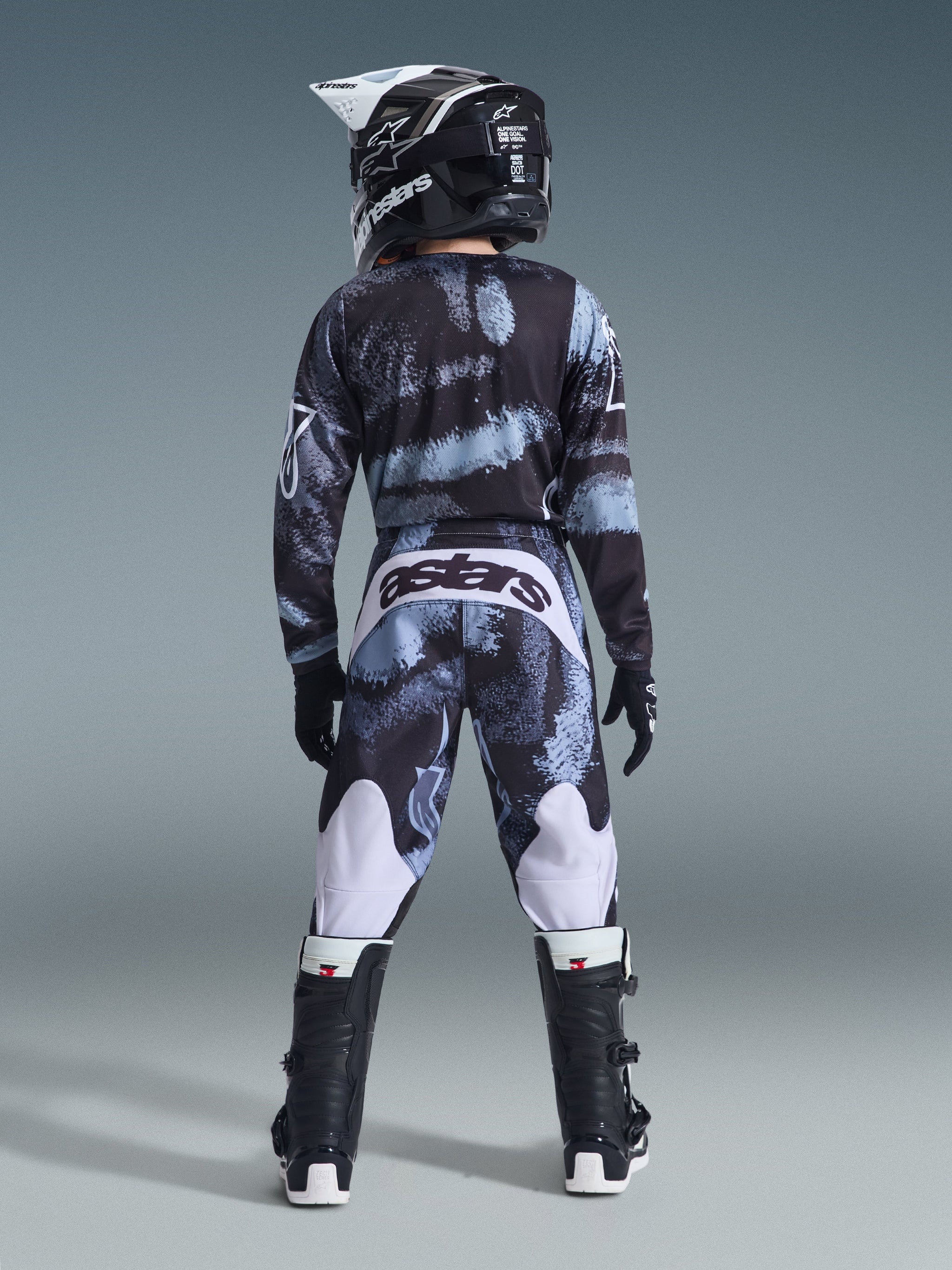 Rear view of a youth rider wearing the Alpinestars 2026 Youth Fluid Lahnd Jersey and matching pants in Iron Camo, featuring a black and grey camouflage pattern with white accents, paired with a black and white helmet and black motocross boots, complete youth off-road racing kit.