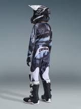 Youth motocross rider wearing the Alpinestars 2026 Youth Fluid Lahnd Jersey and matching pants, black and grey camo pattern with light blue accents, white Astars branding, black helmet with white details, black and white boots, coordinated off-road racing outfit