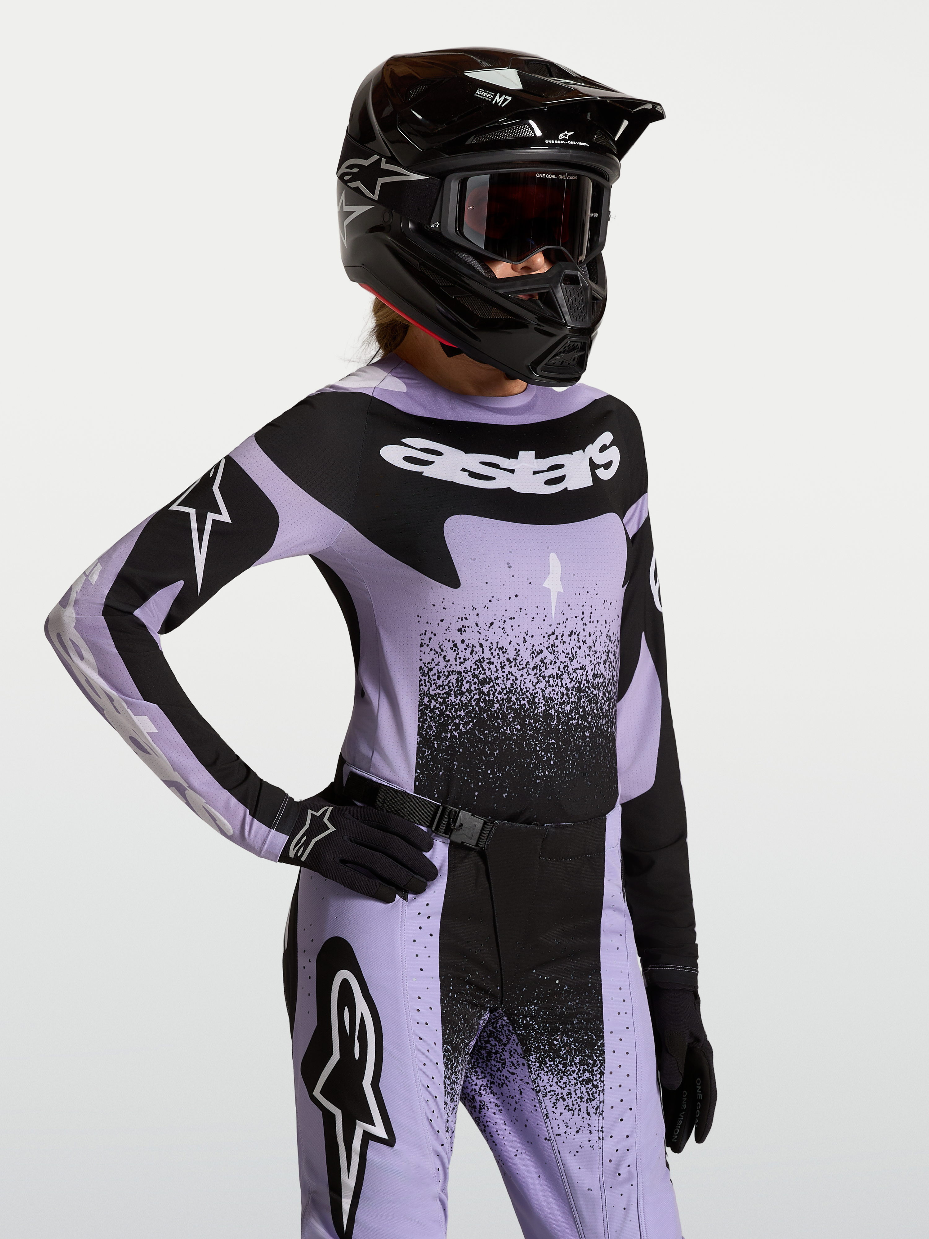 Maillot Alpinestars Stella Techstar Swan, haut de motocross pour femme, violet et noir, coupe athlétique de course, porté par une pilote avec équipement assorti.