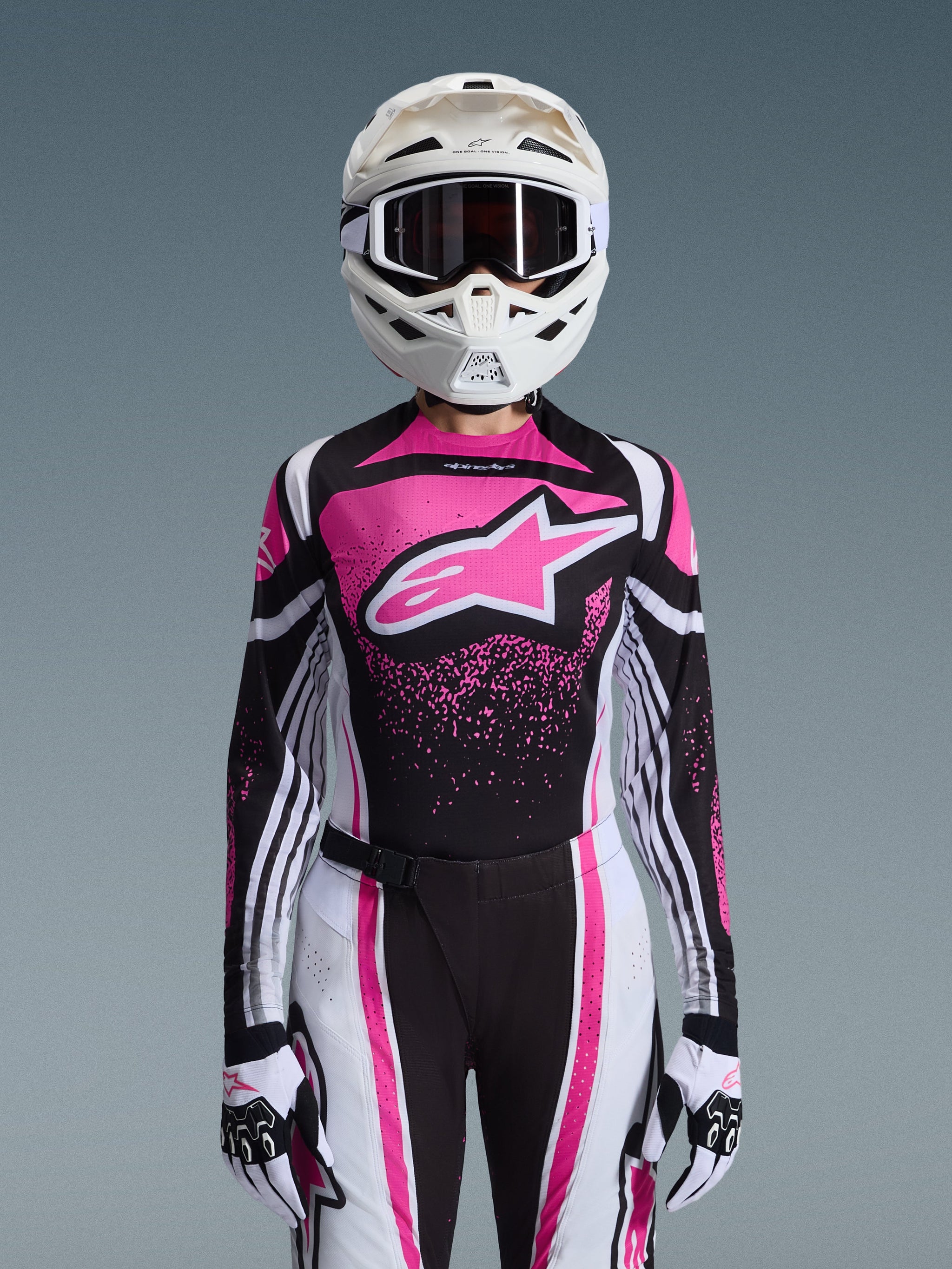 Female motocross rider wearing the 2026 Alpinestars Stella Techstar Nomur Jersey, Black White and Fuchsia design with a large pink logo on the chest, matching racing pants and gloves, white helmet with goggles, complete coordinated gear for off-road competition.