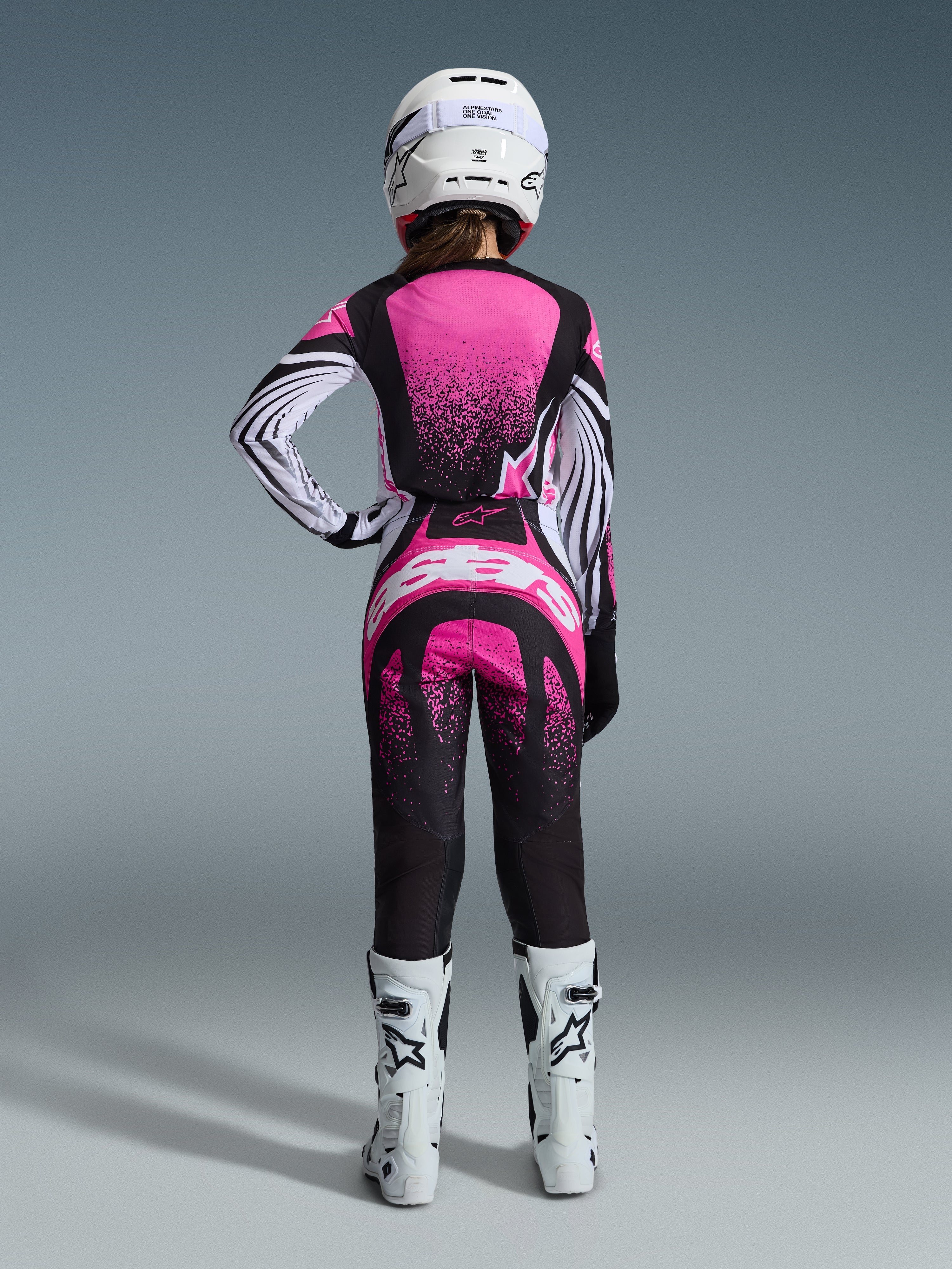 Ensemble d'équipement Alpinestars 2026 Stella Techstar Nomur, pilote de motocross vue de dos, maillot rose et noir avec manches à rayures blanches et motif moucheté, pantalon assorti avec logo Alpinestars blanc, casque blanc et bottes tout-terrain blanches