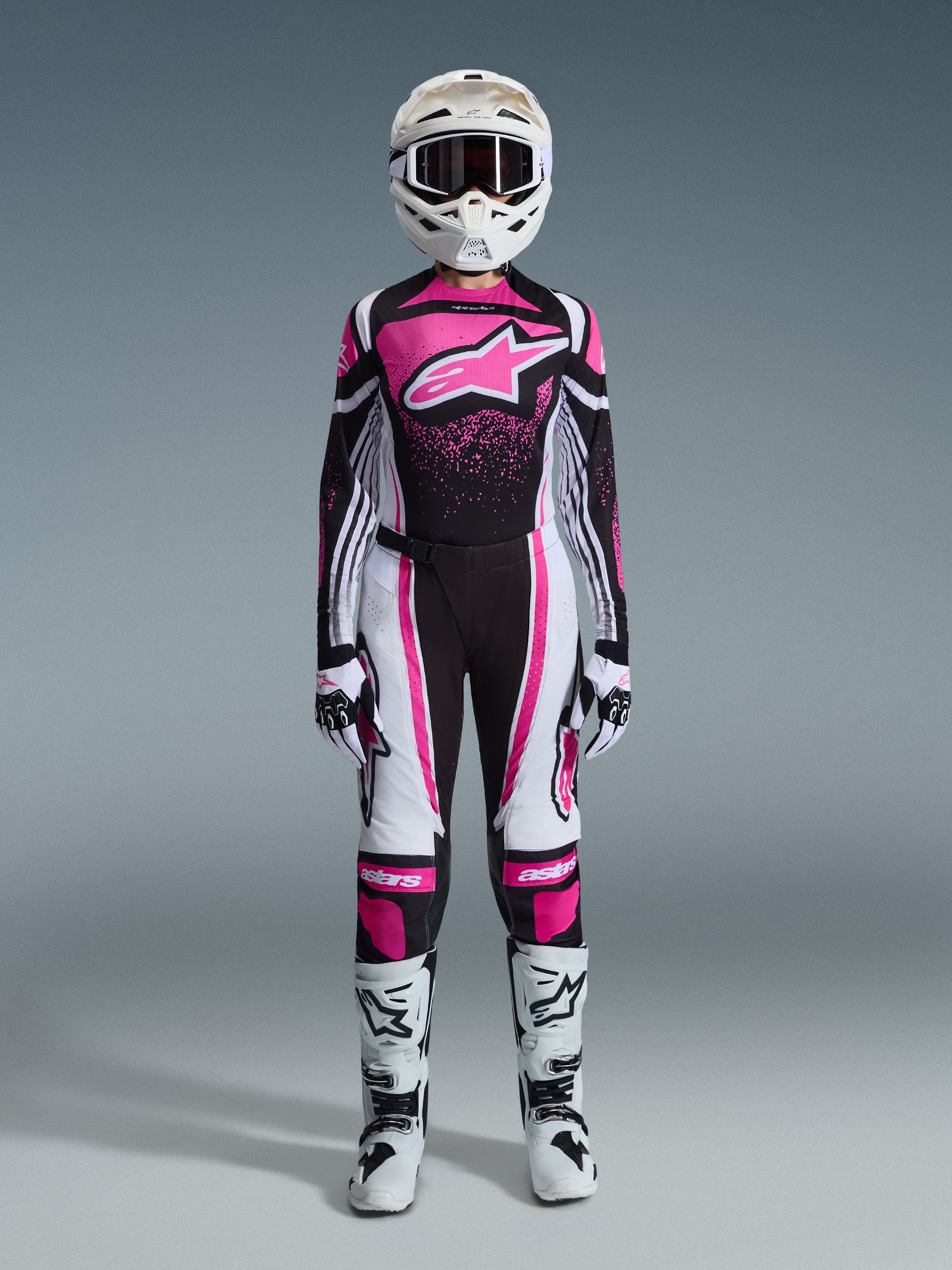 Pilote de motocross femme portant l'ensemble Alpinestars Stella Techstar Nomur 2026, tenue de course rose, noire et blanche, maillot avec un grand logo étoile rose et un motif graphique moucheté, pantalon de performance assorti avec panneaux blancs et roses, casque et lunettes blancs, gants blancs et noirs, et bottes de motocross blanches