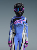Pilote de motocross féminine portant l'ensemble Alpinestars Stella Techstar Nomur 2026, maillot bleu avec logo étoile rose et violet, pantalon bleu assorti avec panneaux latéraux blancs et noirs, casque noir avec lunettes violettes et bleues, gants roses et noirs, tenue de course tout-terrain complète pour femme