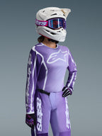 Motocross rider wearing Alpinestars 2026 Woman Stella Fluid Apex Gear Set, Purple and white jersey with Alpinestars logos, matching purple pants, white helmet with purple goggles, and black/purple gloves, professional off-road racing gear