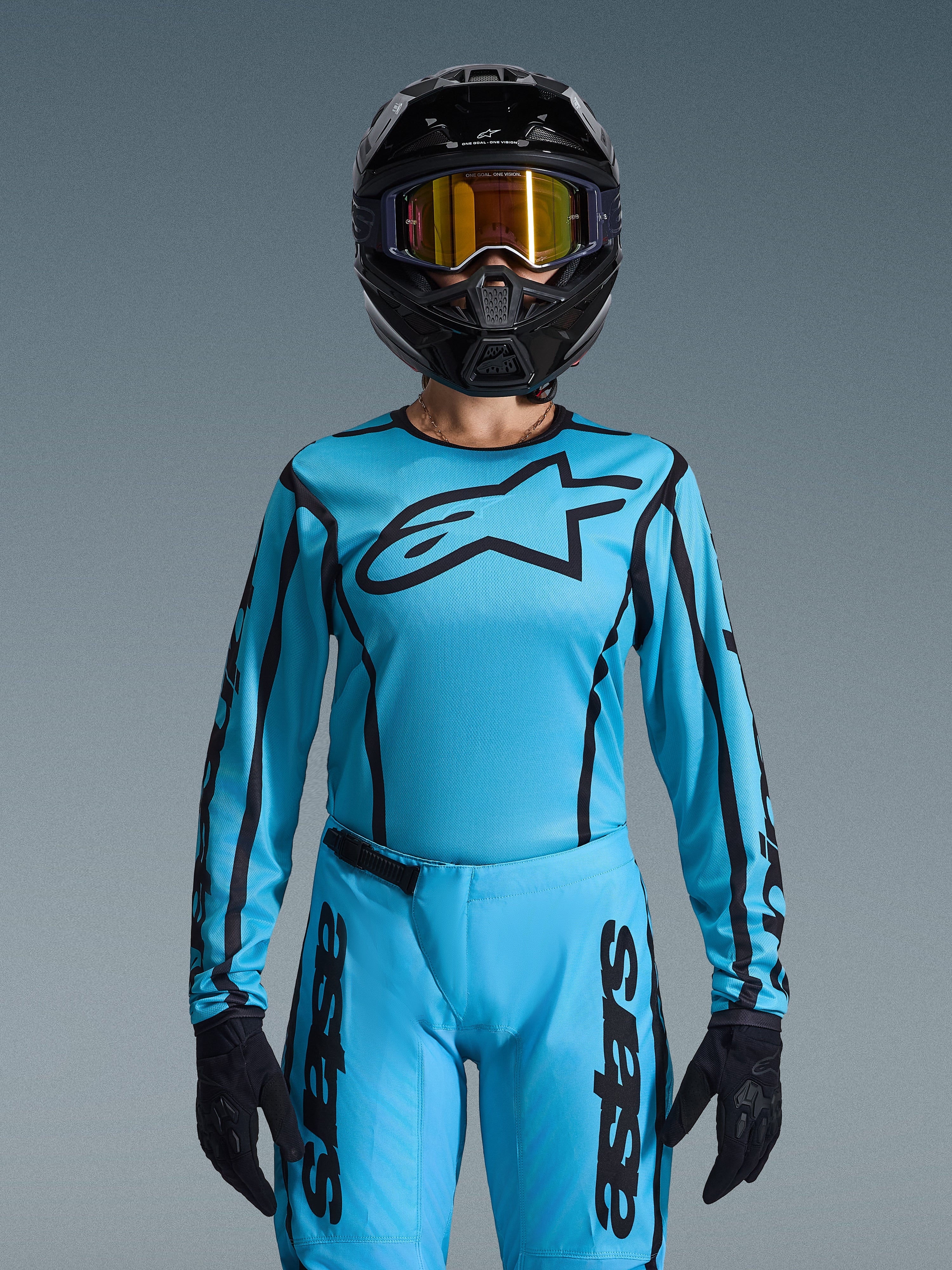 Pilote de motocross portant l'ensemble Stella Fluid Apex Femme 2026 d'Alpinestars, maillot bleu clair avec contours et logo noirs, pantalon assorti, casque noir avec lunettes dorées irisées et gants noirs, tenue de course de motocross coordonnée pour femme