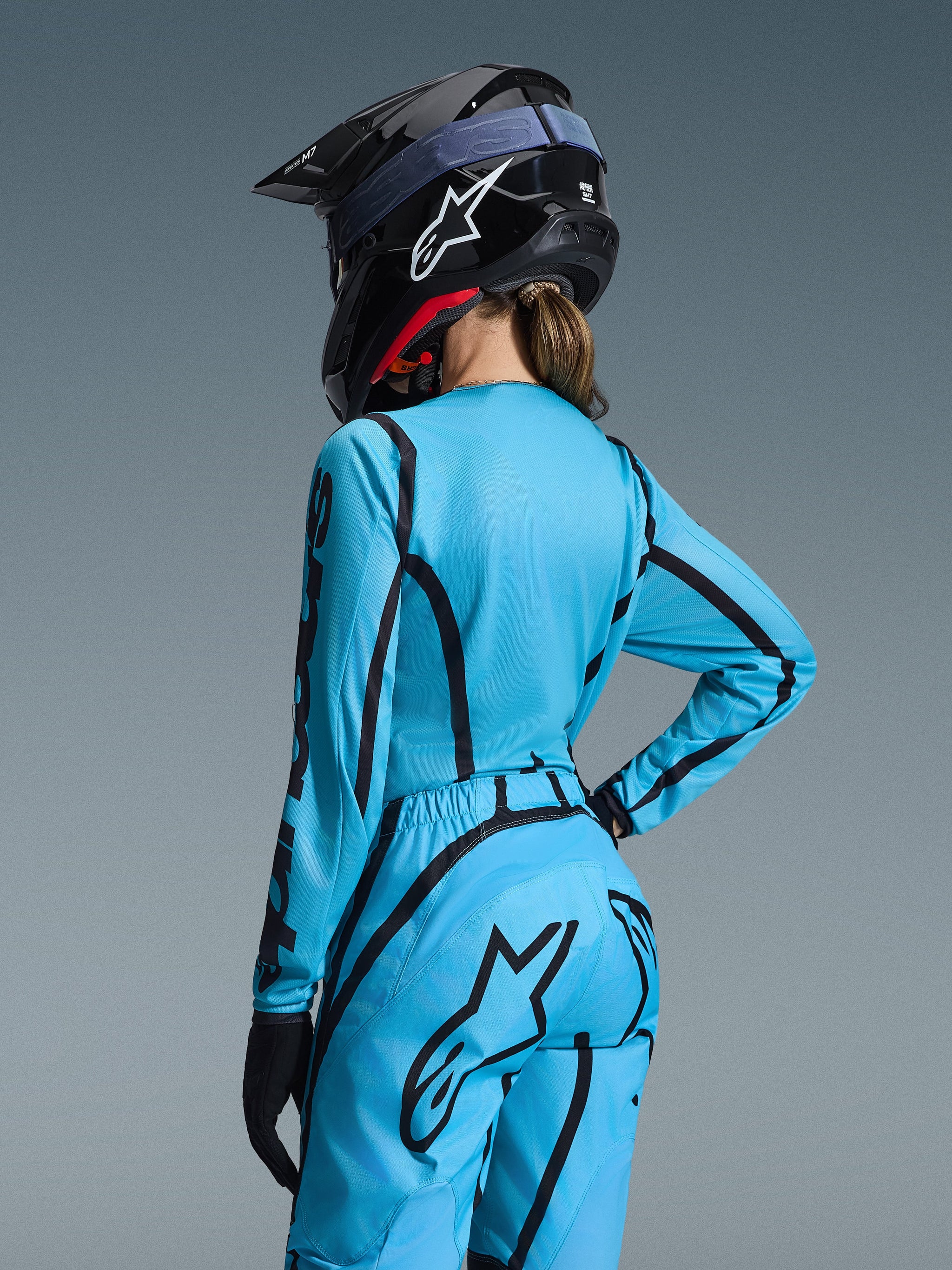 Maillot Stella Fluid Apex Femme 2026 et pantalon assorti, tenue de motocross, bleu clair avec motifs noirs, porté par une pilote vue de dos, avec logo Alpinestars, casque noir et gants noirs, tenue de course tout-terrain professionnelle.