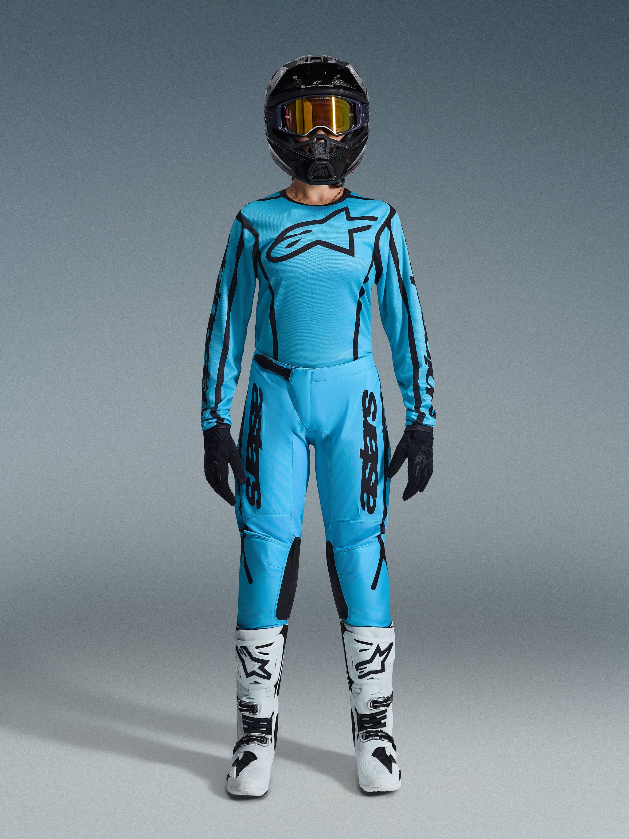 Pilote de motocross portant le maillot Alpinestars Stella Fluid Apex 2026 et le pantalon assorti bleu clair avec des accents noirs, casque noir avec lunettes dorées, bottes blanches et noires, et gants noirs, tenue de motocross complète pour femme.