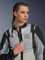 Femme portant la veste Alpinestars Stella ST-2 Air en Ice Gray et noir, portrait en gros plan, avec des panneaux en mesh pour la ventilation, marquage « ALPINESTARS EXPLORATION EQUIPMENT » sur la poitrine avec un logo bleu, veste de moto d'aventure moderne