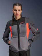 Veste Alpinestars Stella ST-2 Air, veste de moto pour femme, gris foncé et corail fluo, portée par un mannequin aux cheveux tressés, avec panneaux en maille technique pour le flux d'air, perforations laser et marquage « Alpinestars Exploration Equipment » sur la poitrine, équipement de touring d'été
