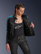 Femme portant la veste Alpinestars Stella T-Dyno Air en Noir Bleu Clair Écru, tenant la veste ouverte pour révéler la doublure intérieure en maille et la poche imperméable, associée à un débardeur noir à logo et un pantalon de moto noir, équipement de moto urbain d'inspiration vintage.