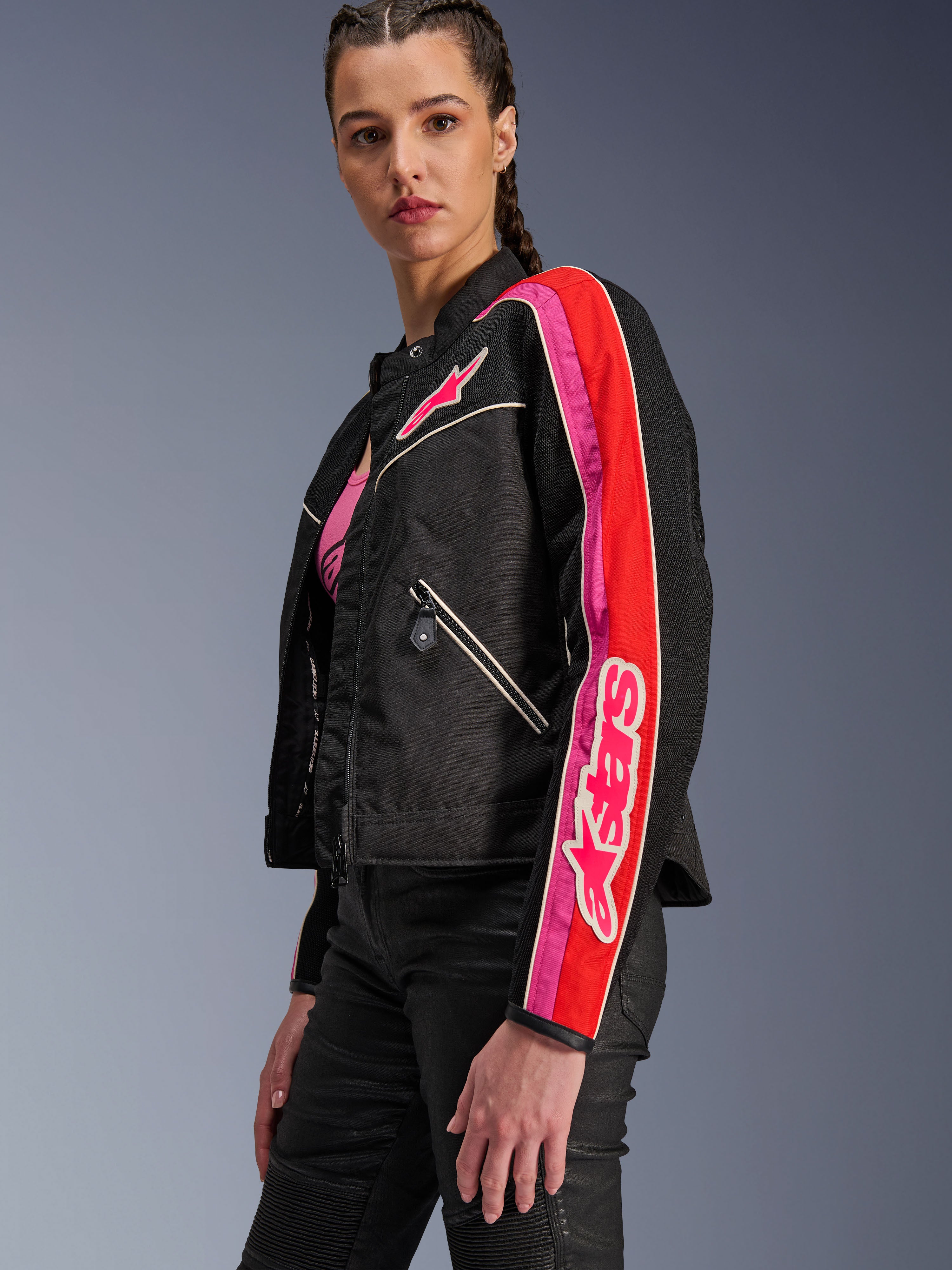 Veste Alpinestars Stella T-Dyno Air pour femme, veste de moto pour femme, noire avec accents rouge vif et fuchsia, portée avec un pantalon noir, arborant le logo Astars rose sur la poitrine et la manche, coupe biker ajustée avec panneaux en maille pour la ventilation.