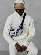 Sweat à capuche Alpinestars Essentials Ageless, streetwear décontracté, blanc cassé, petit logo noir brodé sur la poitrine, capuche portée sur une casquette beige avec marquage ASTARS, stylisé avec un sac bandoulière gris clair, vêtement lifestyle premium