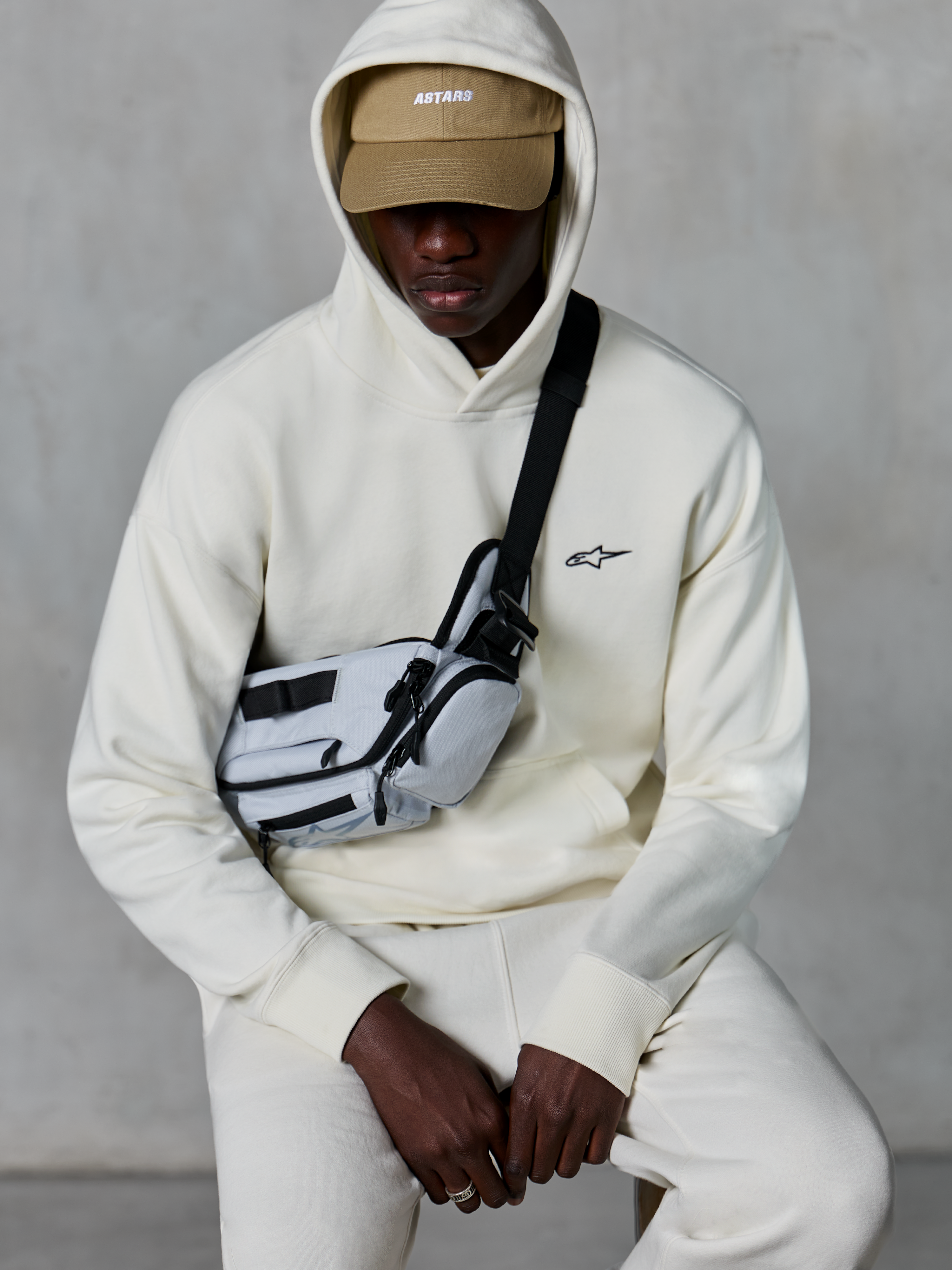 Sweat à capuche Alpinestars Essentials Ageless, streetwear décontracté, blanc cassé, petit logo noir brodé sur la poitrine, capuche portée sur une casquette beige avec marquage ASTARS, stylisé avec un sac bandoulière gris clair, vêtement lifestyle premium