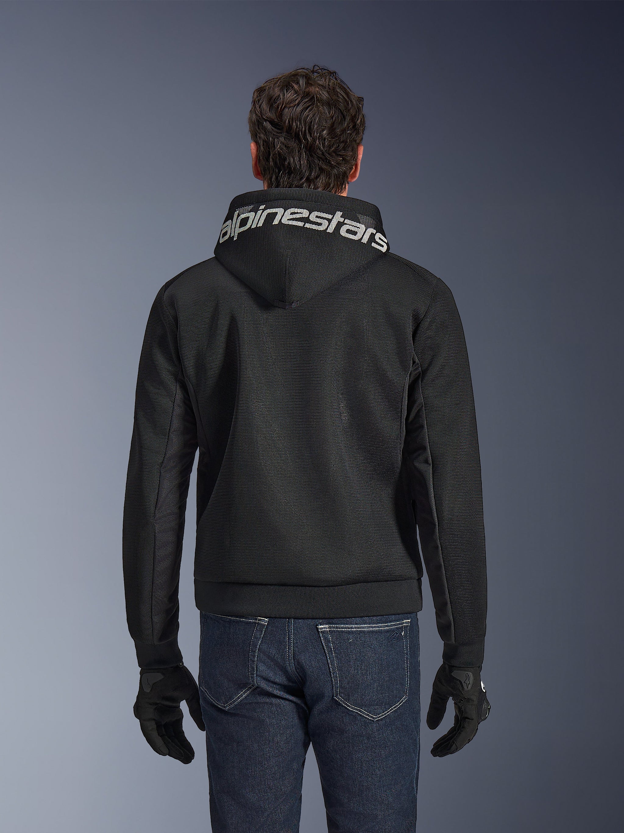 Alpinestars Chrome Superairflow Hoodie, Black motorcycle hoodie, rear view featuring bold white Alpinestars branding across the hood, 3D technical mesh construction, worn with dark blue jeans and black gloves, ventilated urban riding apparel