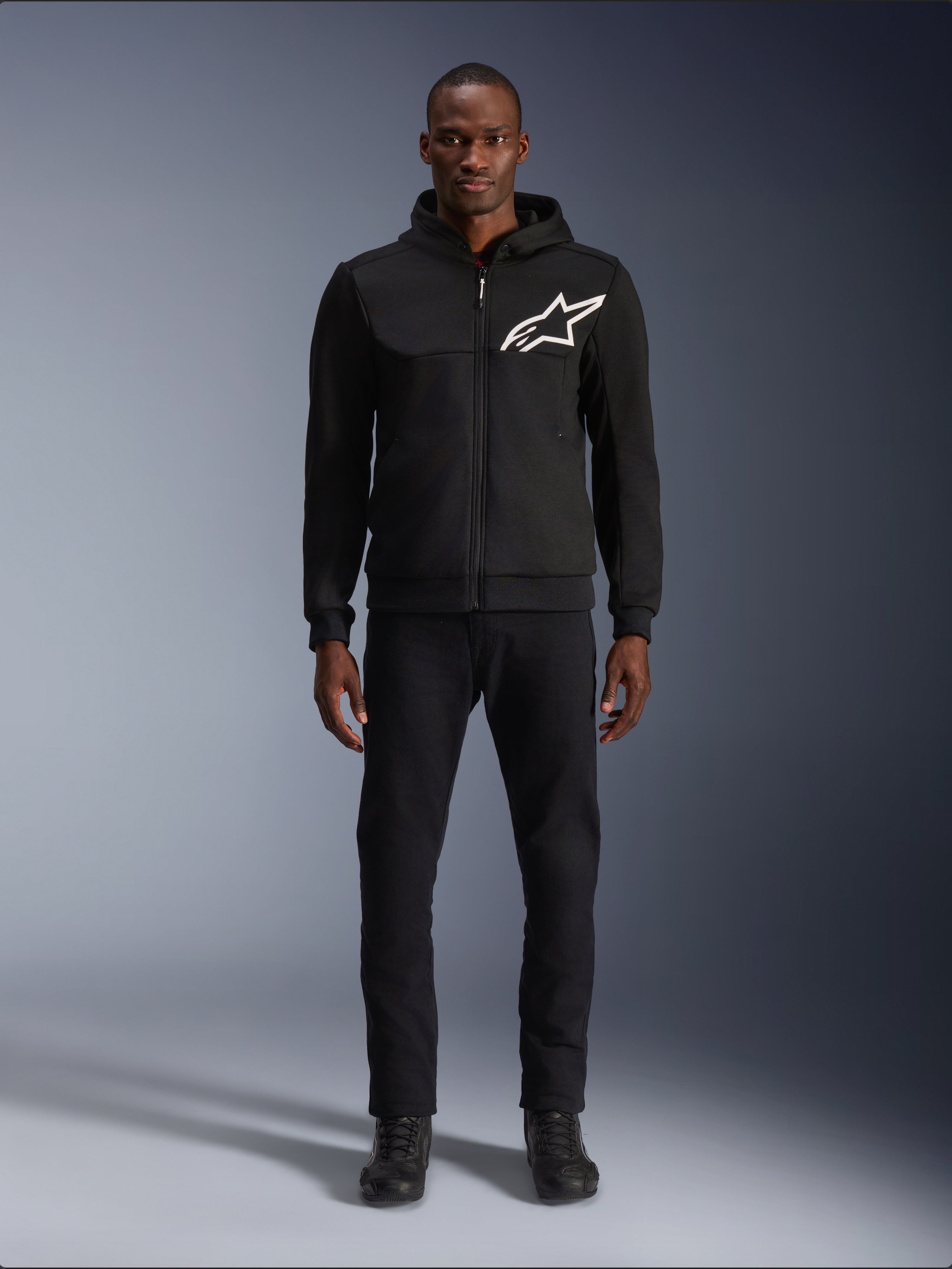 Alpinestars Chrome V2 Sport Hoodie, Black and White motorcycle hoodie, featuring a large white Alpinestars logo on the chest, zip-up front and hood, worn by a model standing with black pants and boots, urban-style protective riding gear.