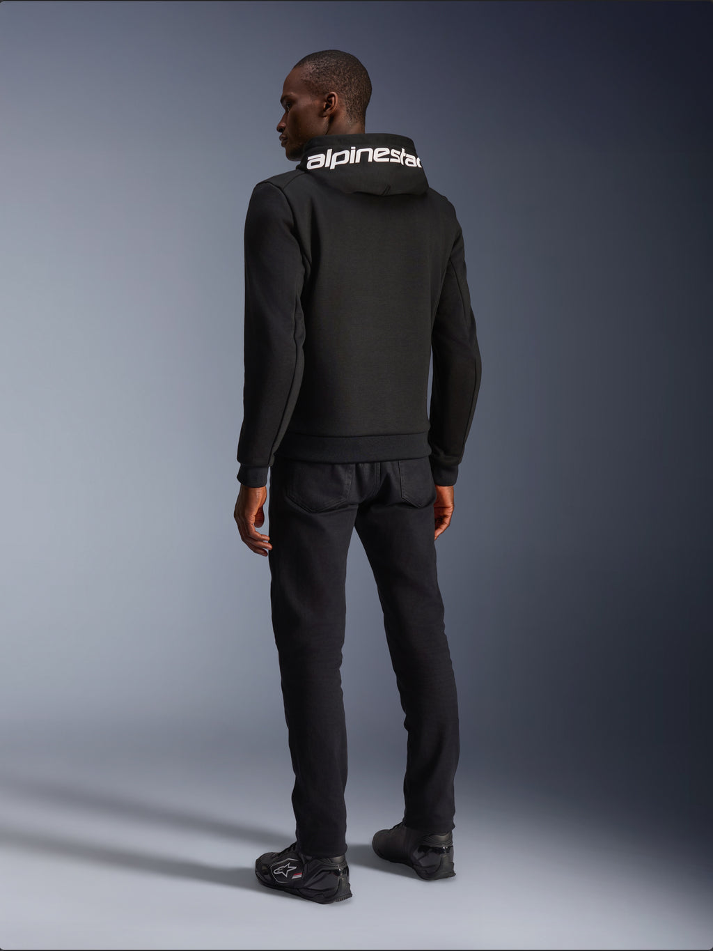 Alpinestars Chrome V2 Sport Hoodie, Black, rear view showing white Alpinestars branding on the hood, worn with black pants and riding shoes, casual urban motorcycle apparel with an athletic silhouette