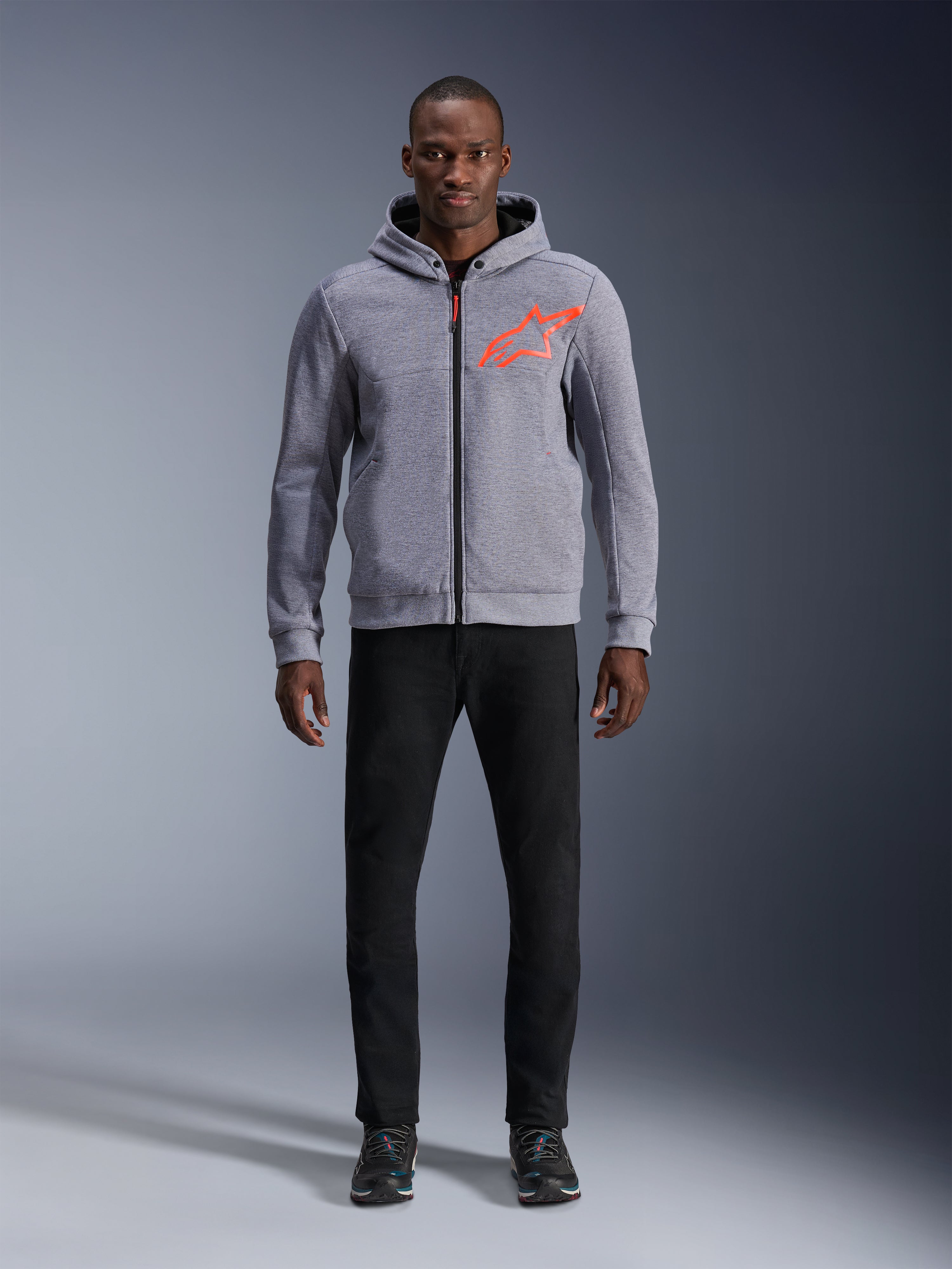Model wearing Alpinestars Chrome V2 Sport Hoodie in Gray Heather Red Fluo, light gray tech fleece hoodie with a bright orange-red star logo on the chest, paired with black pants and dark sneakers, casual urban motorcycle riding style