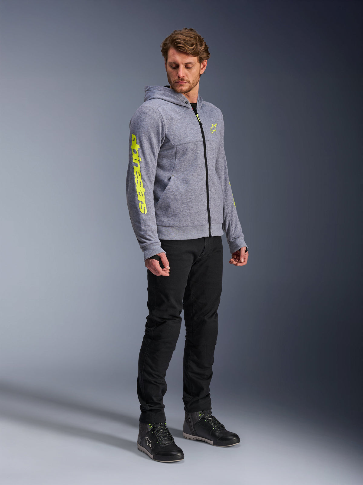 Alpinestars Chrome V2 Sport Hoodie, gris chiné et jaune fluo, porté par un mannequin homme avec un pantalon noir et des chaussures de moto noires, avec un logo jaune vif sur la manche et la poitrine, équipement de moto urbain et décontracté