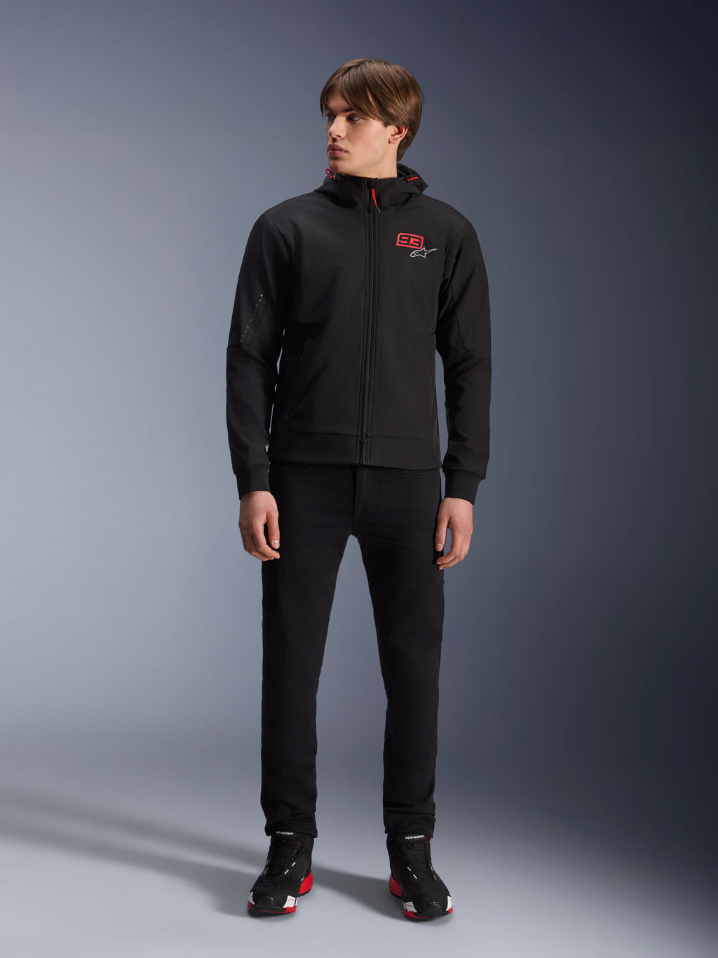 Sweat à capuche Alpinestars MM93 Radium Tech, sweat à capuche de moto urbain noir, doté d'un logo 93 rouge et d'un logo étoilé blanc sur la poitrine, porté par un mannequin avec un pantalon noir et des chaussures de moto assorties, coque technique coupe-vent élégante pour la conduite urbaine.