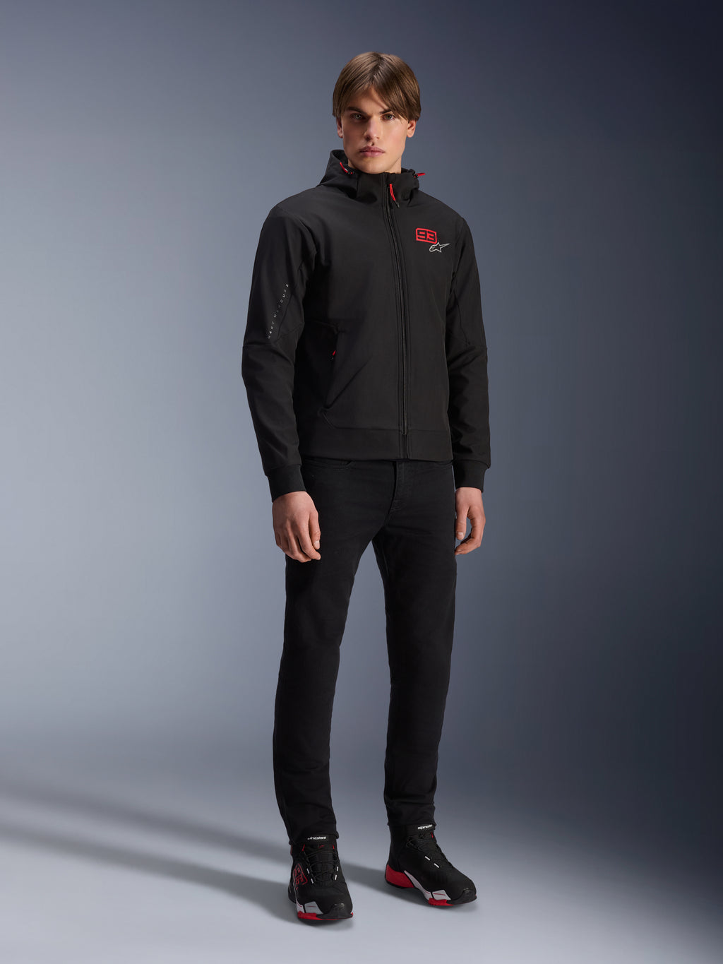 Sweat à capuche Alpinestars MM93 Radium Tech, veste de moto urbaine, noir avec des touches de rouge vif, porté par un mannequin avec un pantalon noir et des chaussures de moto, présentant le logo '93' rouge sur la poitrine et le marquage Marc Marquez sur la manche, streetwear technique décontracté pour la conduite urbaine