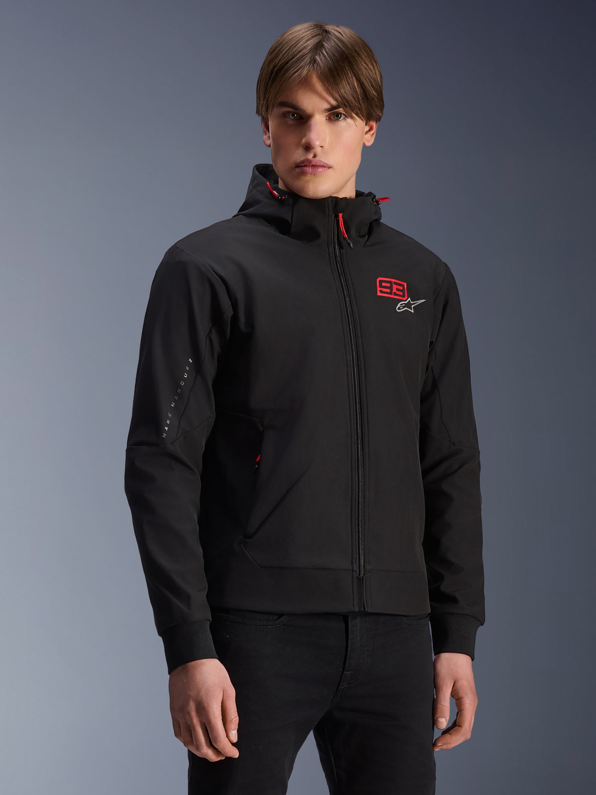 MM93 Radium Tech Hoodie, veste de moto urbaine zippée noire, numéro 93 rouge et logo Alpinestars blanc sur la poitrine, porté par un mannequin avec un pantalon noir, doté d'une capuche amovible avec cordons de serrage rouges et marquage sur la manche, design décontracté inspiré du sport automobile