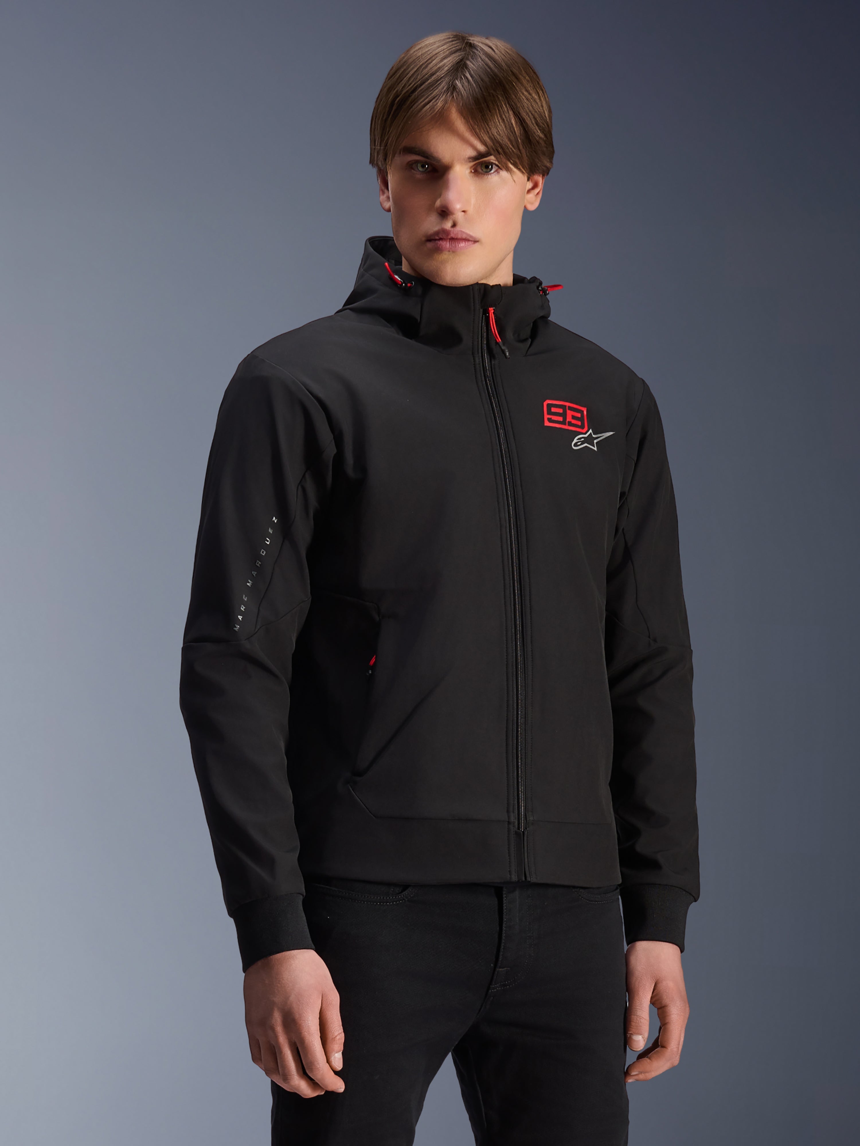 MM93 Radium Tech Hoodie, black urban motorcycle zip-up jacket, red 93 and white Alpinestars logo on chest, worn by model with black pants, featuring a removable hood with red drawstrings and sleeve branding, casual motorsport-inspired design