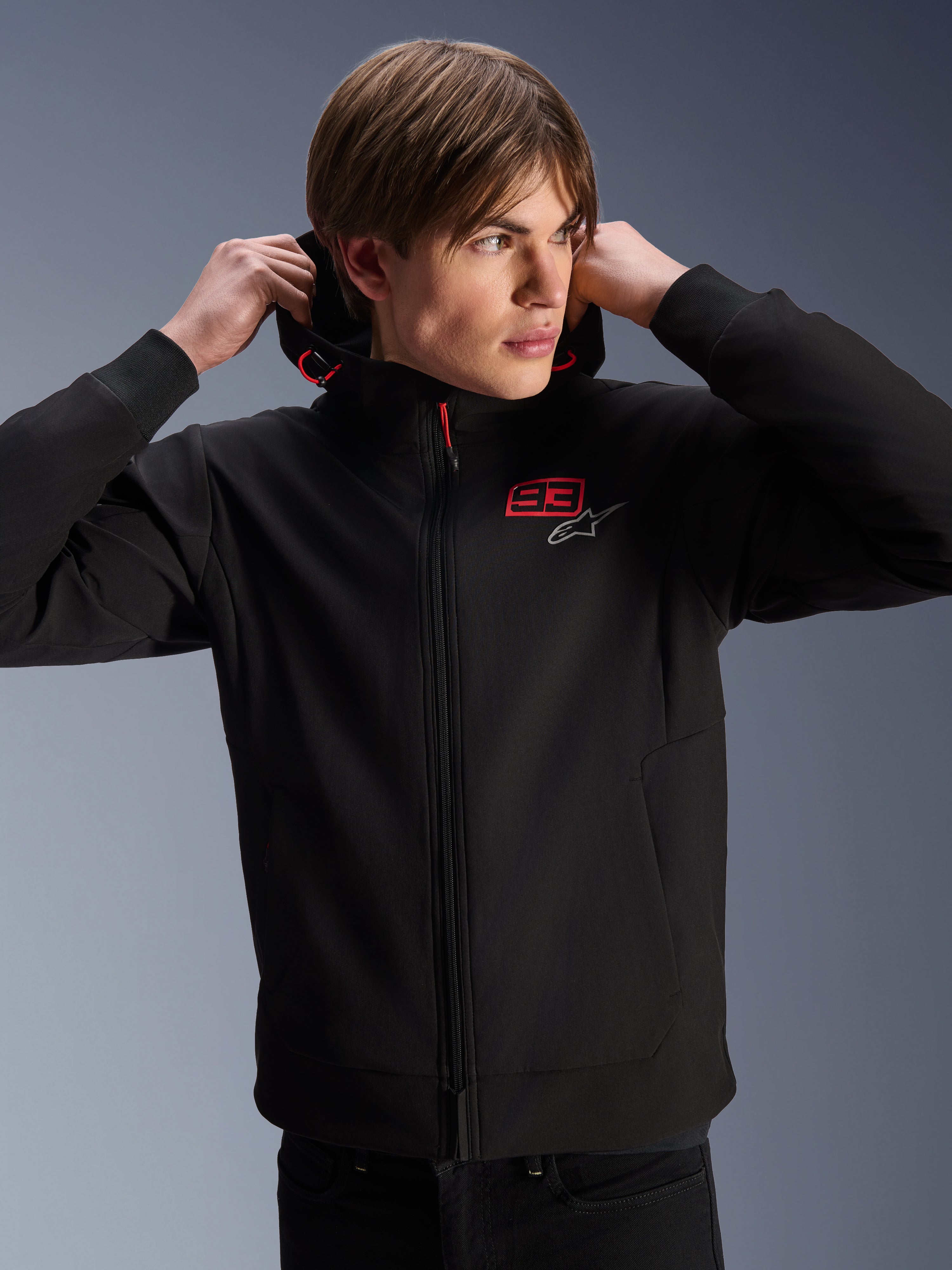 Sweat à capuche Alpinestars MM93 Radium Tech, veste de moto urbaine, noir avec logos rouges et argentés, porté par un mannequin homme, doté d'une fermeture éclair intégrale et de poignets côtelés, vêtement de sport automobile décontracté