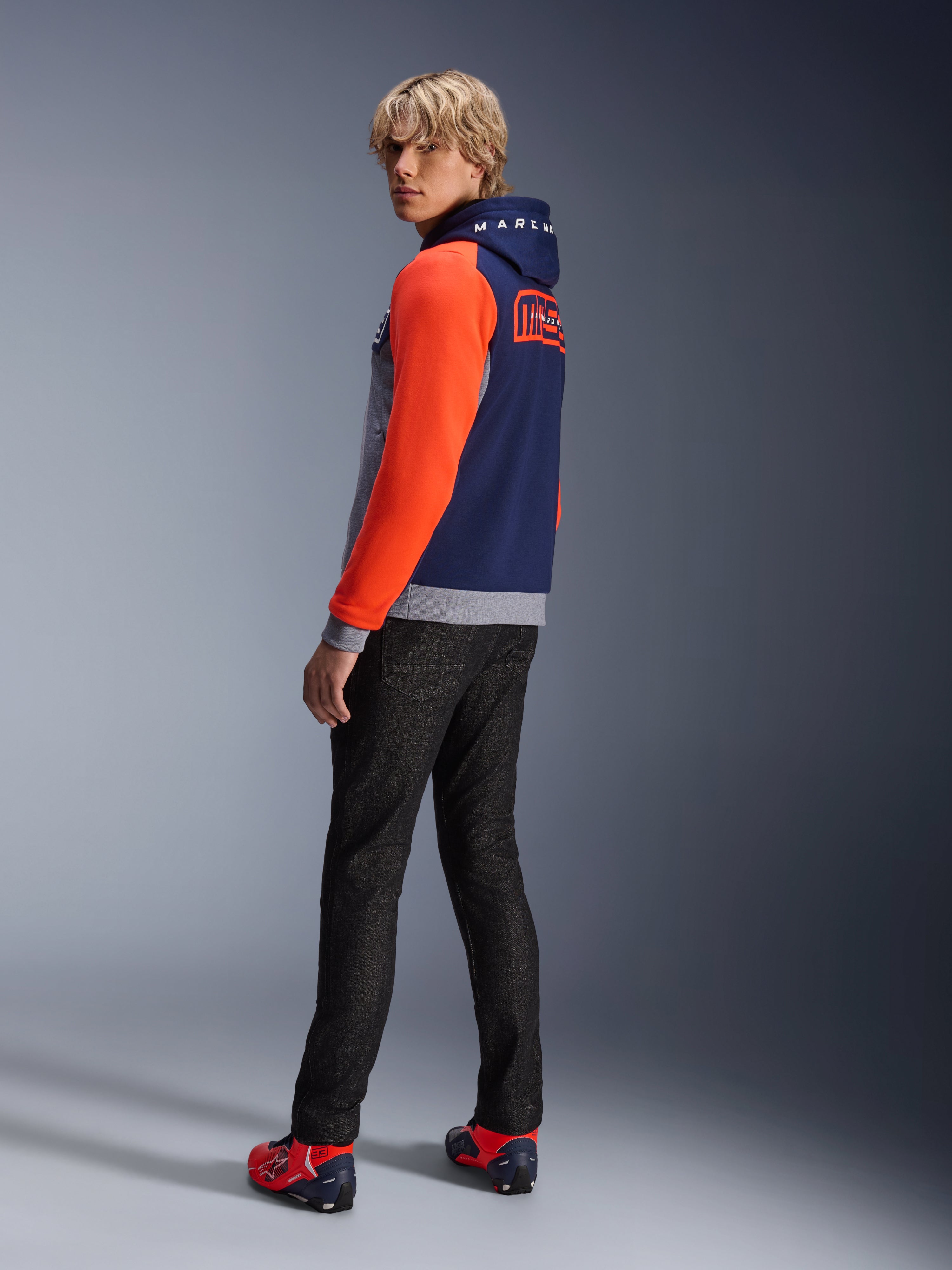Alpinestars MM93 Chrome V2 Sport Hoodie, Blue Navy Gray Mel Red Fluo, model wearing hoodie with bright red sleeves and MM93 Marc Marquez branding on the back, styled with dark jeans and red riding shoes, casual urban motorcycle gear