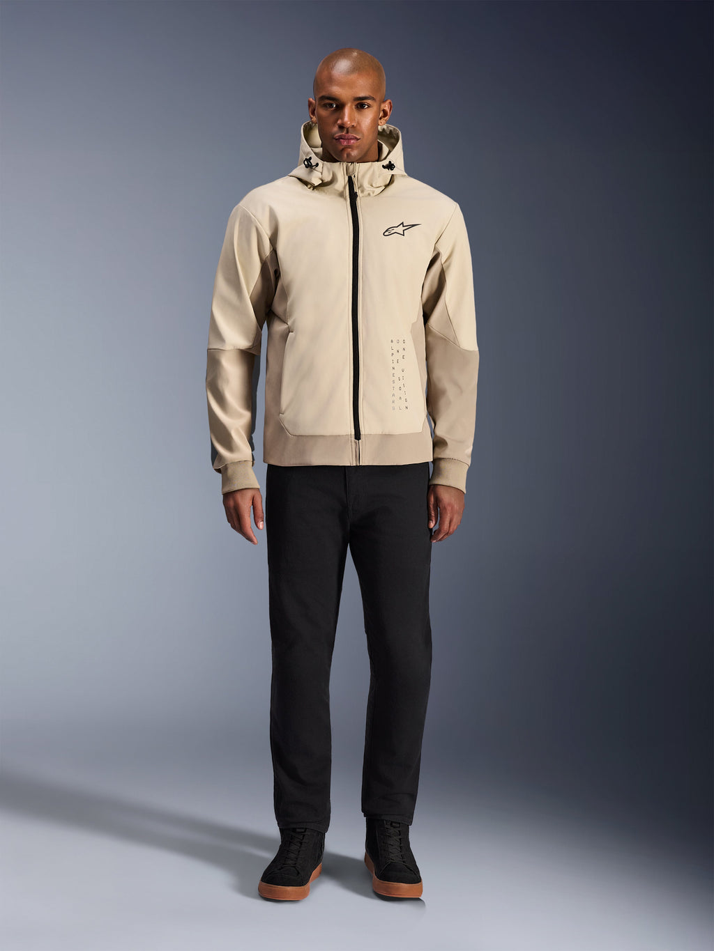 Sweat à capuche Alpinestars Radium Tech en Mastic Aluminum, hoodie de moto urbain porté par un mannequin homme avec un pantalon noir et des bottes noires, doté d'une fermeture éclair noire intégrale, d'un petit logo sur la poitrine et d'une coupe athlétique pour la conduite urbaine.