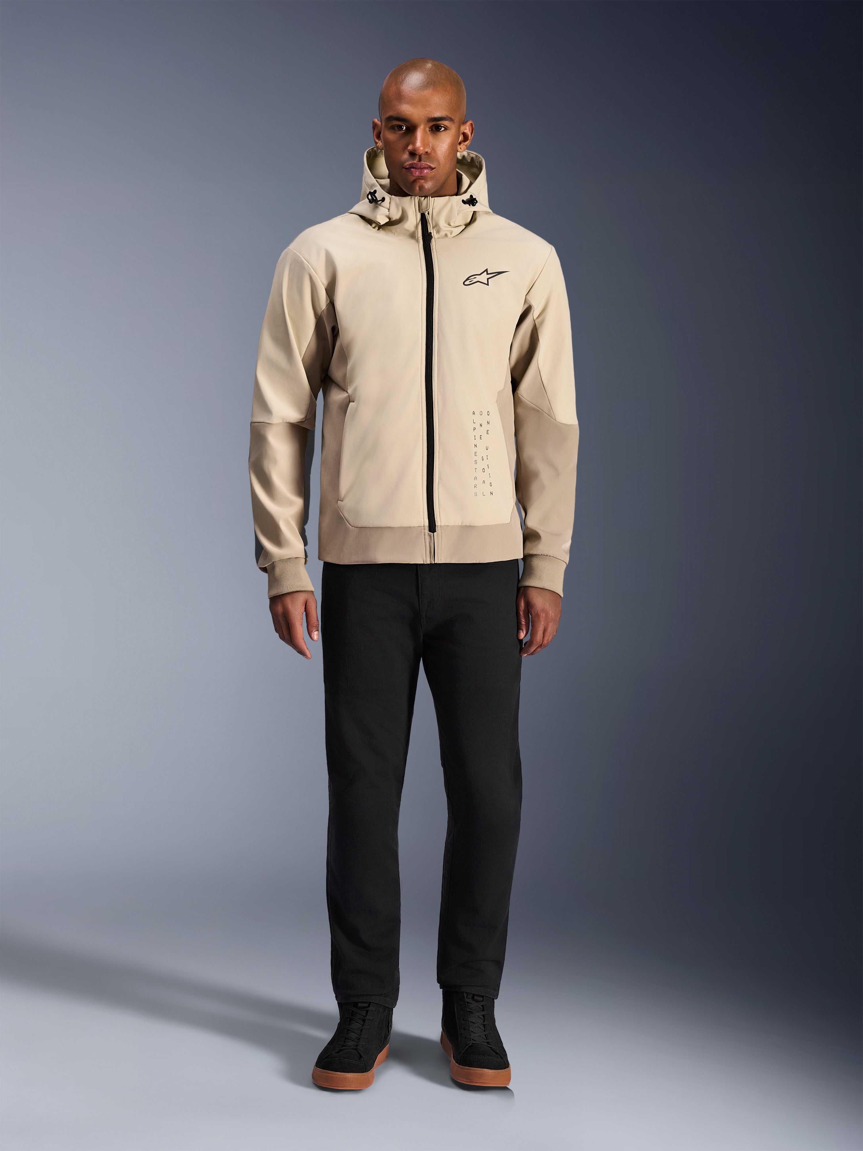 Sweat à capuche Alpinestars Radium Tech en Mastic Aluminum, hoodie de moto urbain porté par un mannequin homme avec un pantalon noir et des bottes noires, doté d'une fermeture éclair noire intégrale, d'un petit logo sur la poitrine et d'une coupe athlétique pour la conduite urbaine.