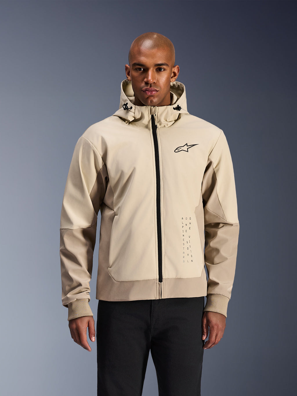 Alpinestars Radium Tech Hoodie en Mastic Aluminum, veste de moto urbaine beige clair et brun clair, fermeture éclair frontale noire et petit logo sur la poitrine, portée par un homme en pantalon noir, coque technique coupe-vent au style streetwear contemporain