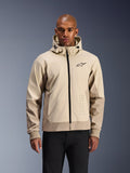Alpinestars Radium Tech Hoodie en Mastic Aluminum, veste de moto urbaine beige clair et brun clair, fermeture éclair frontale noire et petit logo sur la poitrine, portée par un homme en pantalon noir, coque technique coupe-vent au style streetwear contemporain