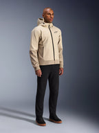 Alpinestars Radium Tech Hoodie, veste de moto urbaine, Mastic Aluminum beige clair, porté avec un pantalon noir et des baskets montantes noires, doté d'une coque technique coupe-vent, fermeture éclair frontale et capuche amovible, design streetwear moderne pour la conduite urbaine
