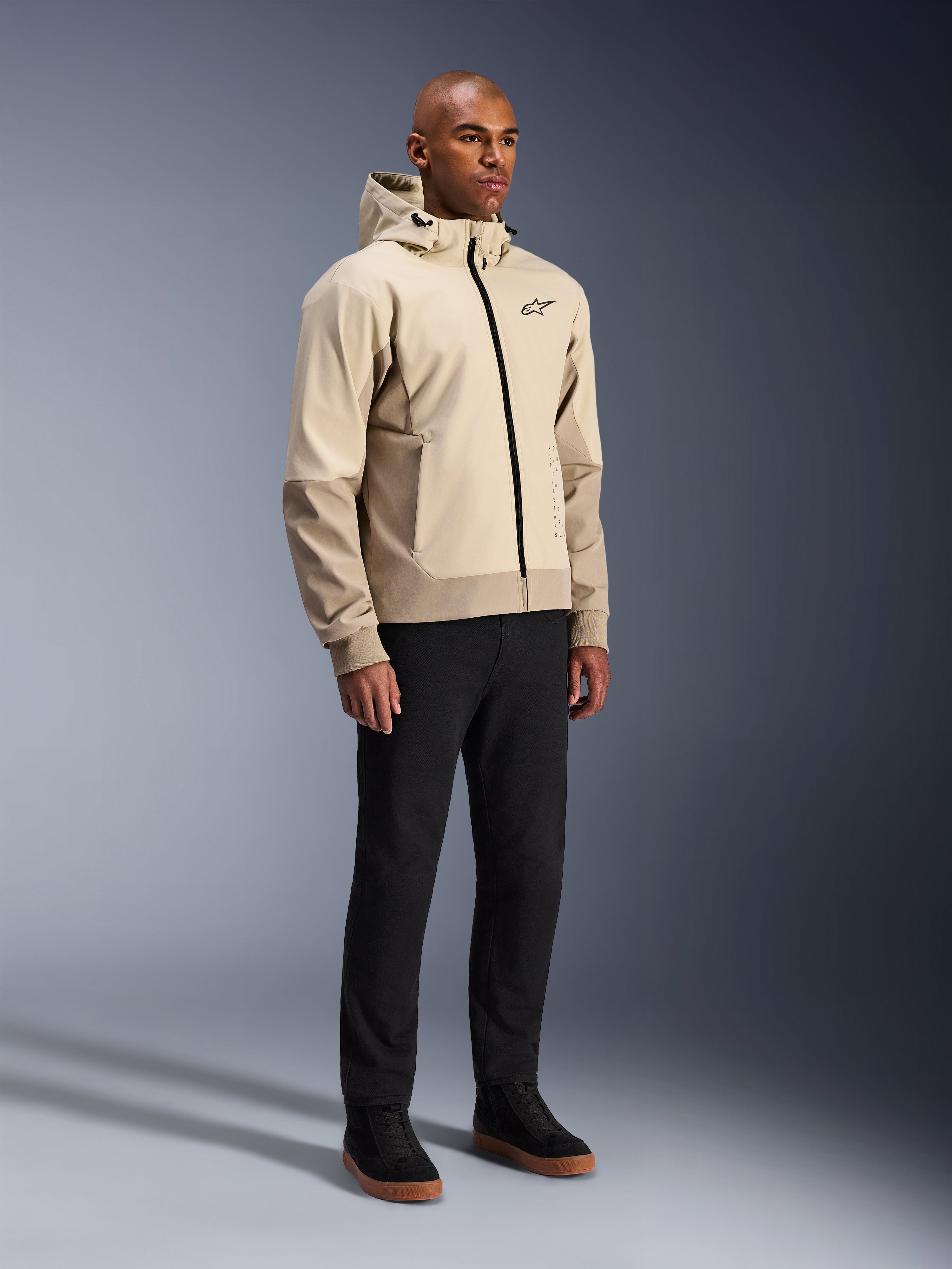 Alpinestars Radium Tech Hoodie, veste de moto urbaine, Mastic Aluminum beige clair, porté avec un pantalon noir et des baskets montantes noires, doté d'une coque technique coupe-vent, fermeture éclair frontale et capuche amovible, design streetwear moderne pour la conduite urbaine