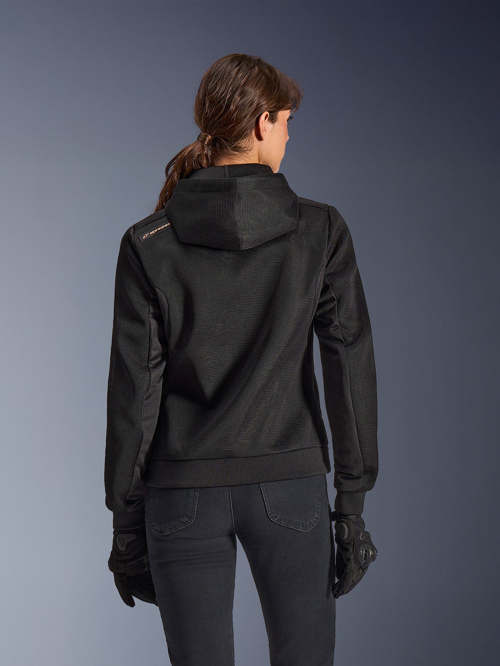 Alpinestars Stella Chrome Superairflow Hoodie, vue de dos d'une femme portant un sweat à capuche de moto technique en maille 3D noire, avec capuche technique et logo cuivré sur l'épaule, porté avec des gants et un pantalon de moto noirs.