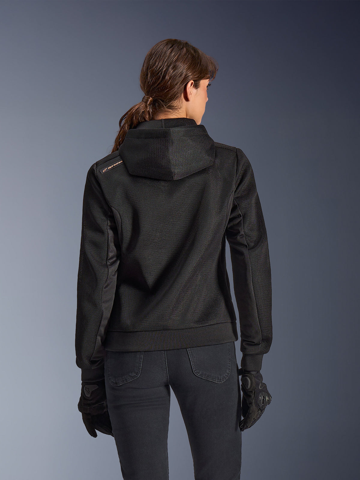 Alpinestars Stella Chrome Superairflow Hoodie, vue de dos d'une femme portant un sweat à capuche de moto technique en maille 3D noire, avec capuche technique et logo cuivré sur l'épaule, porté avec des gants et un pantalon de moto noirs.