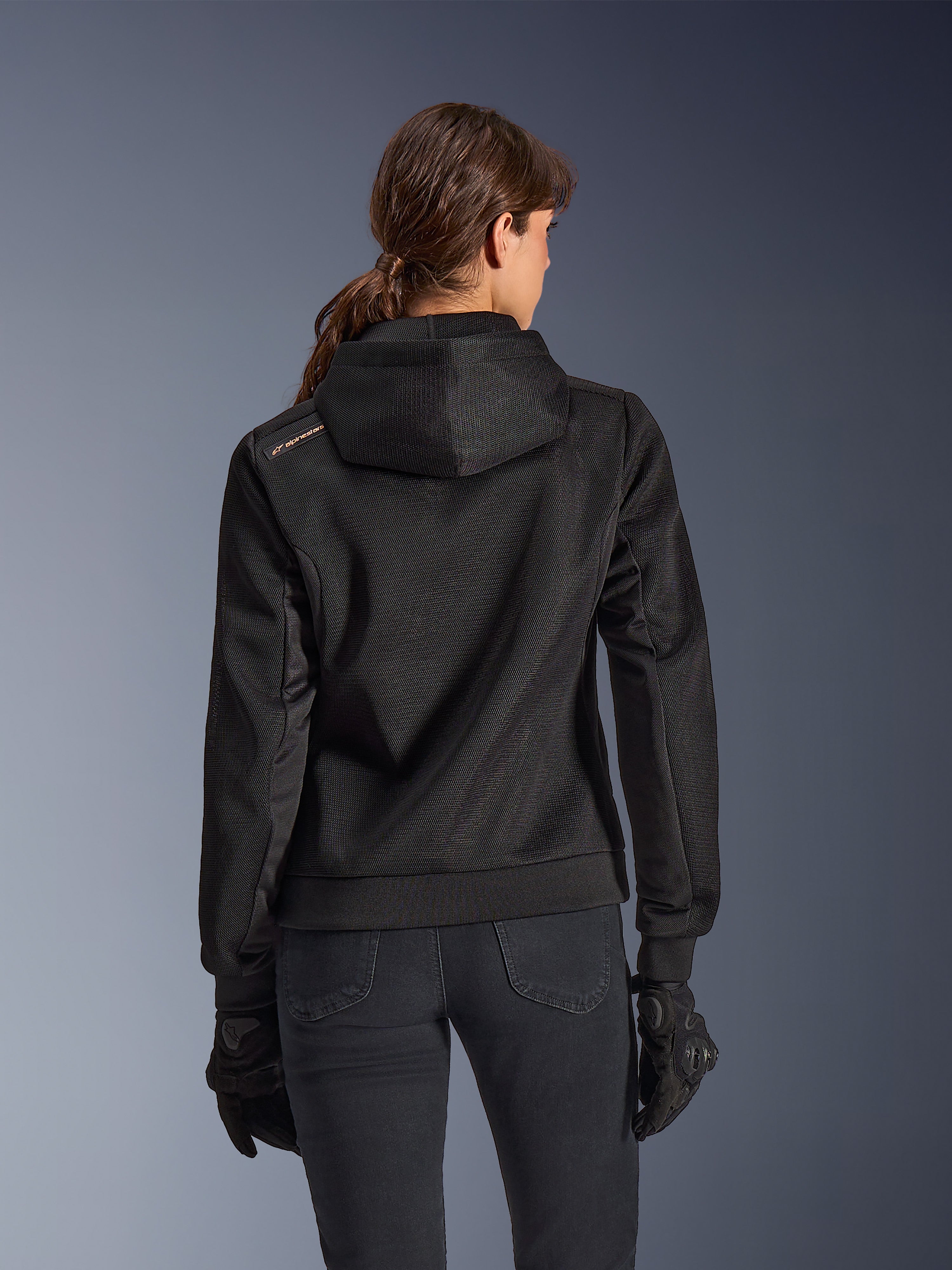 Alpinestars Stella Chrome Superairflow Hoodie, vue de dos d'une femme portant un sweat à capuche de moto technique en maille 3D noire, avec capuche technique et logo cuivré sur l'épaule, porté avec des gants et un pantalon de moto noirs.